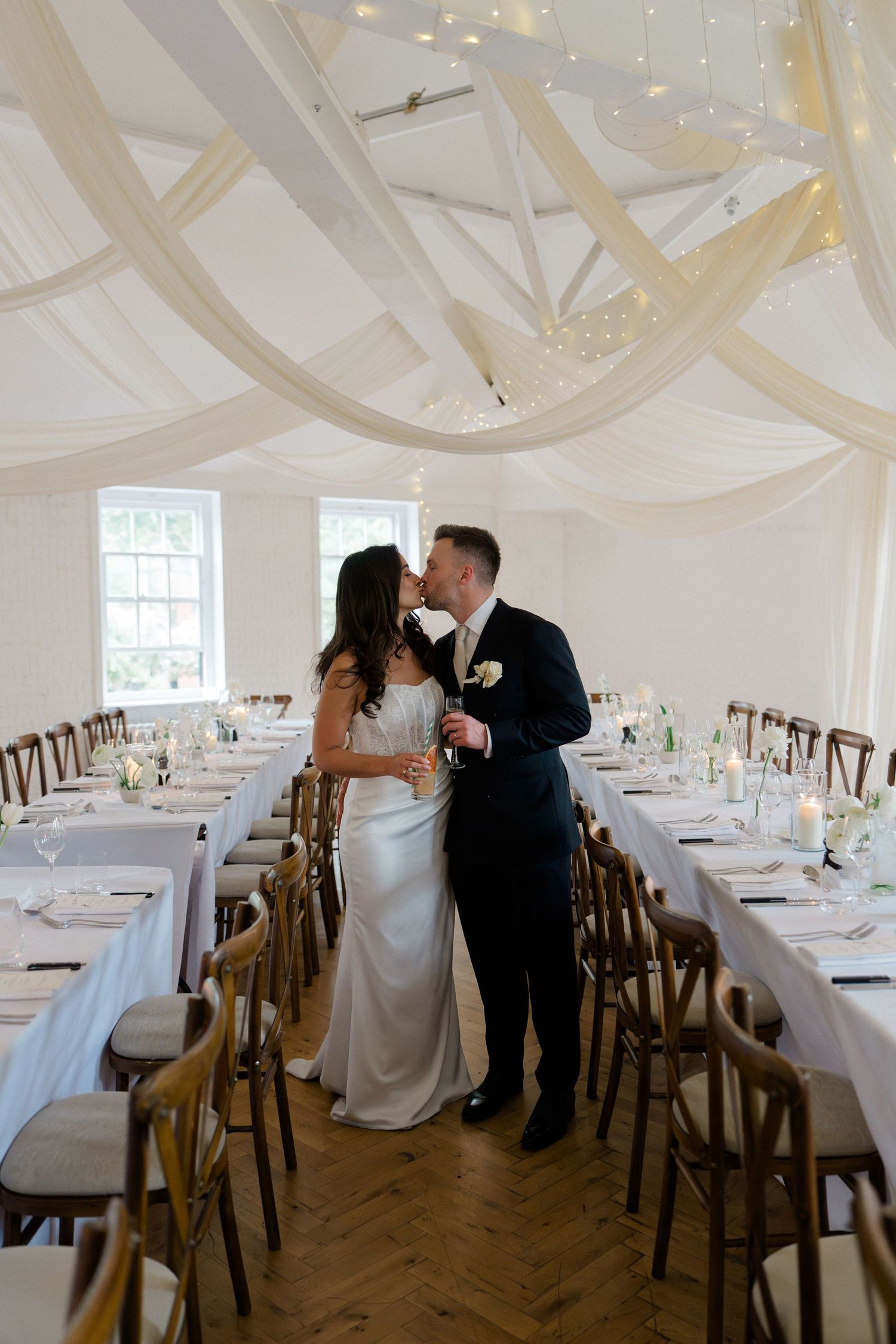 Bride and groom kissing in elegant white draped wedding reception hall with long decorated tables.