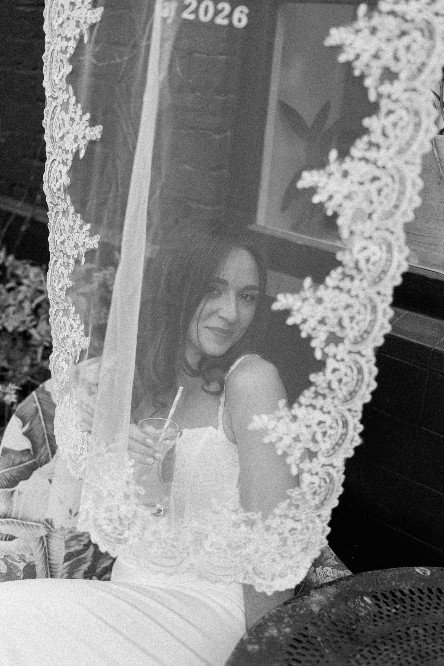 Black and white photo of a smiling bride holding a drink framed by a lace wedding veil.