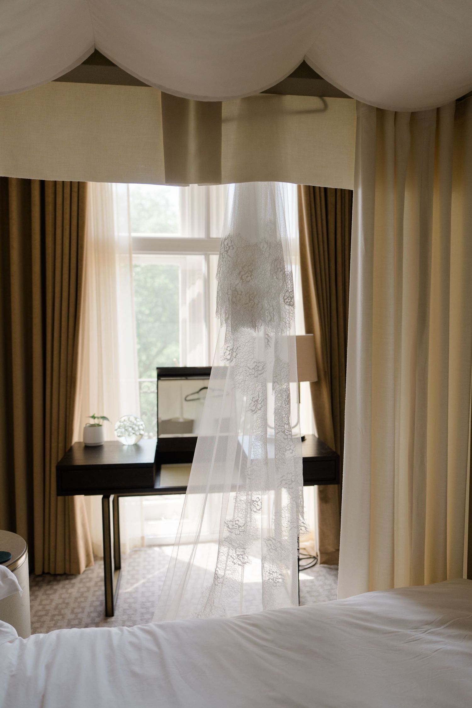Bridal veil hanging near a window in an elegant hotel room with gold curtains and a vanity desk.