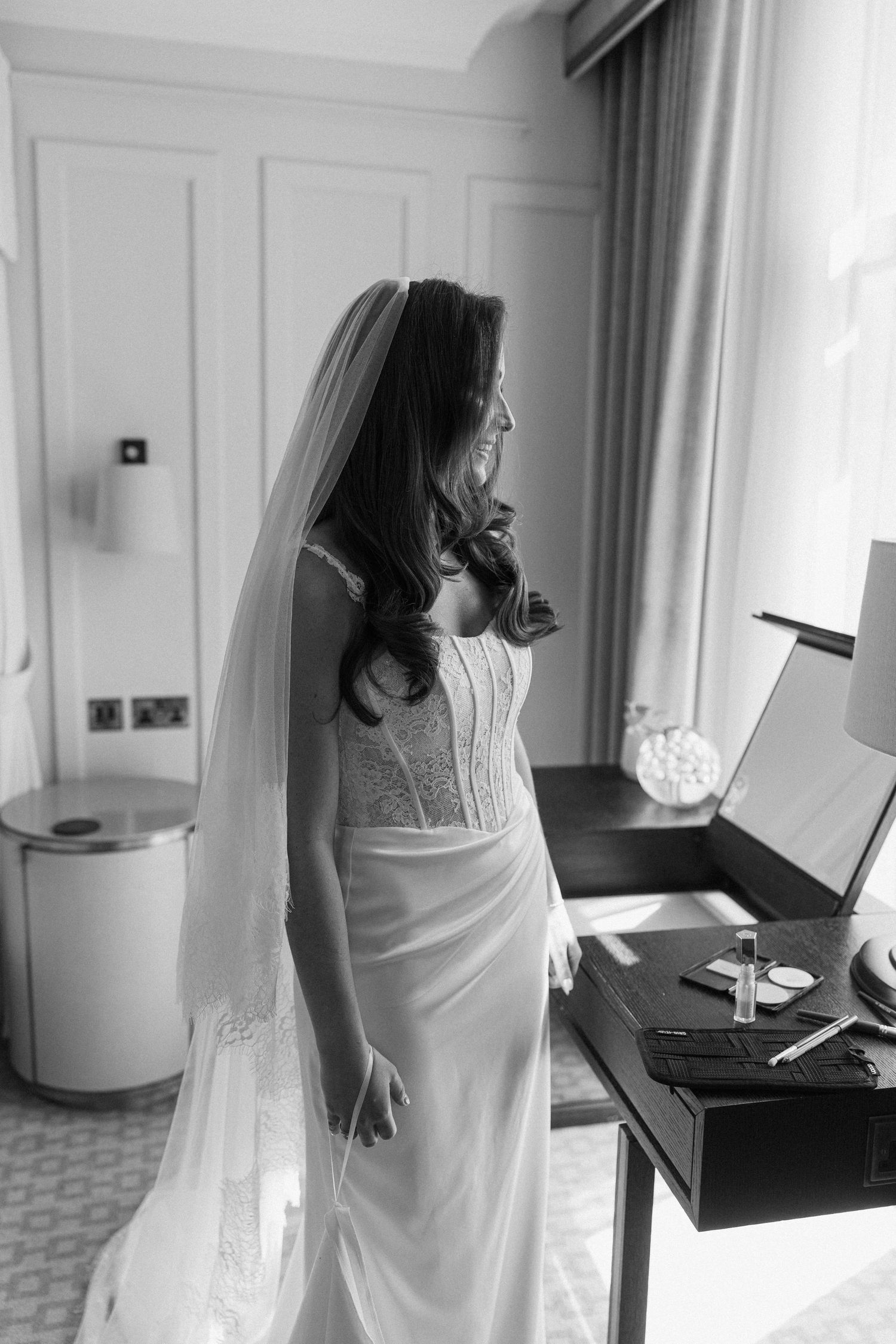 Elegant black and white portrait of a dark-haired bride in a lace wedding gown and veil standing by a window.