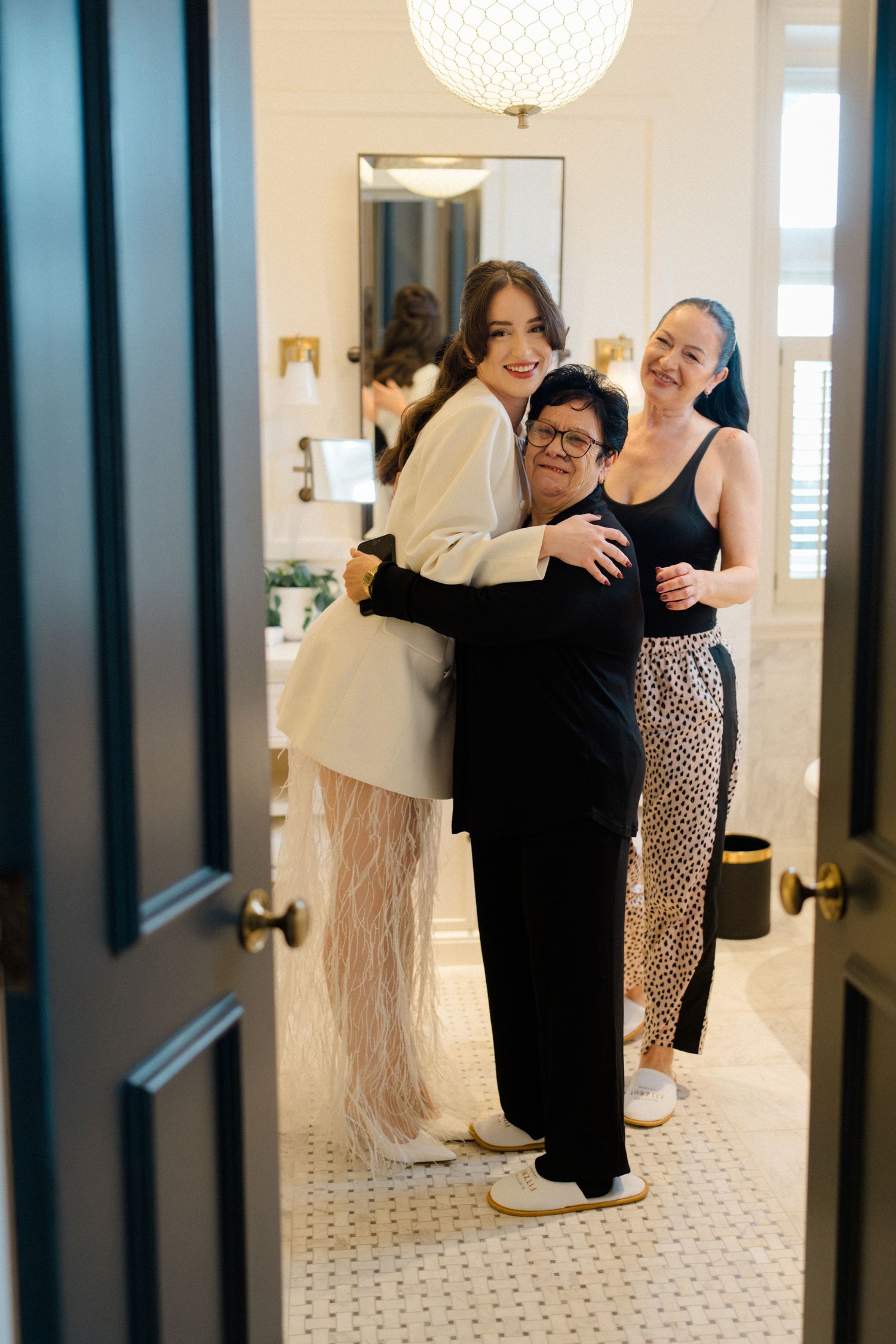 Three women embrace and smile together in an elegant white marble bathroom during wedding preparations.