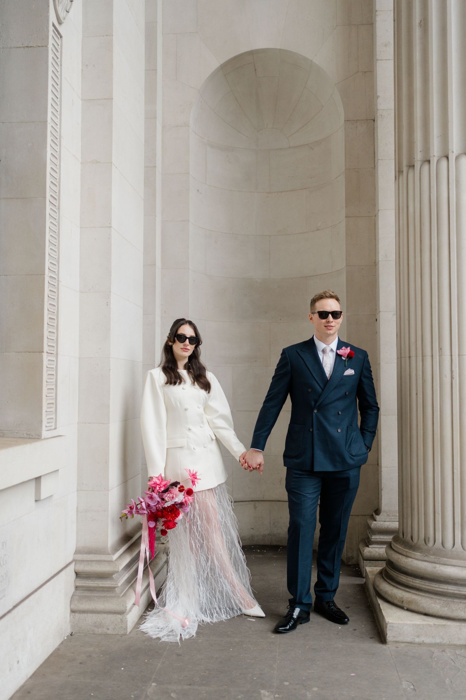 Bride and groom holding hands between stone columns, bride in white feathered skirt suit with bouquet.