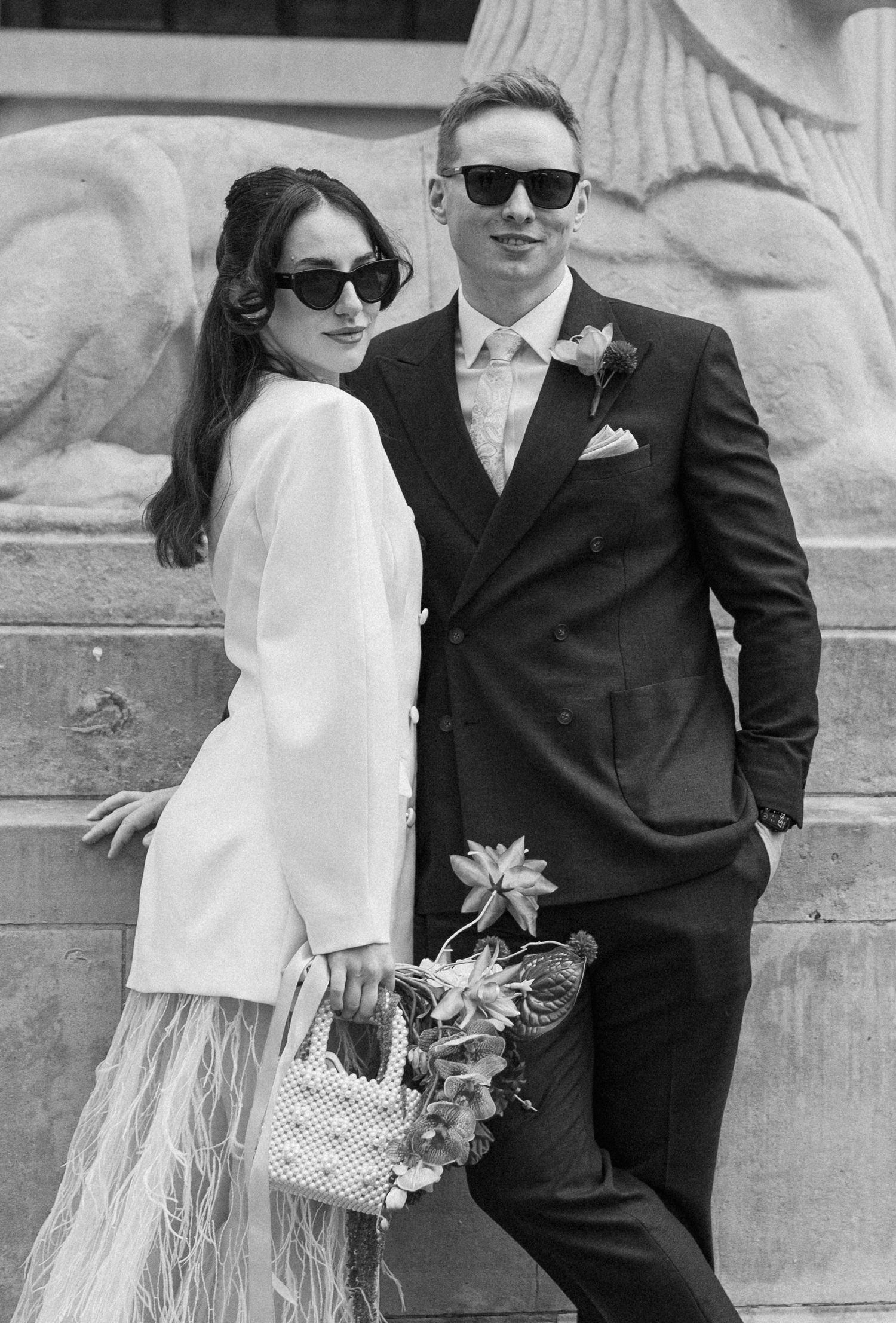 Stylish bride and groom in sunglasses posing together in black and white wedding portrait outdoors.