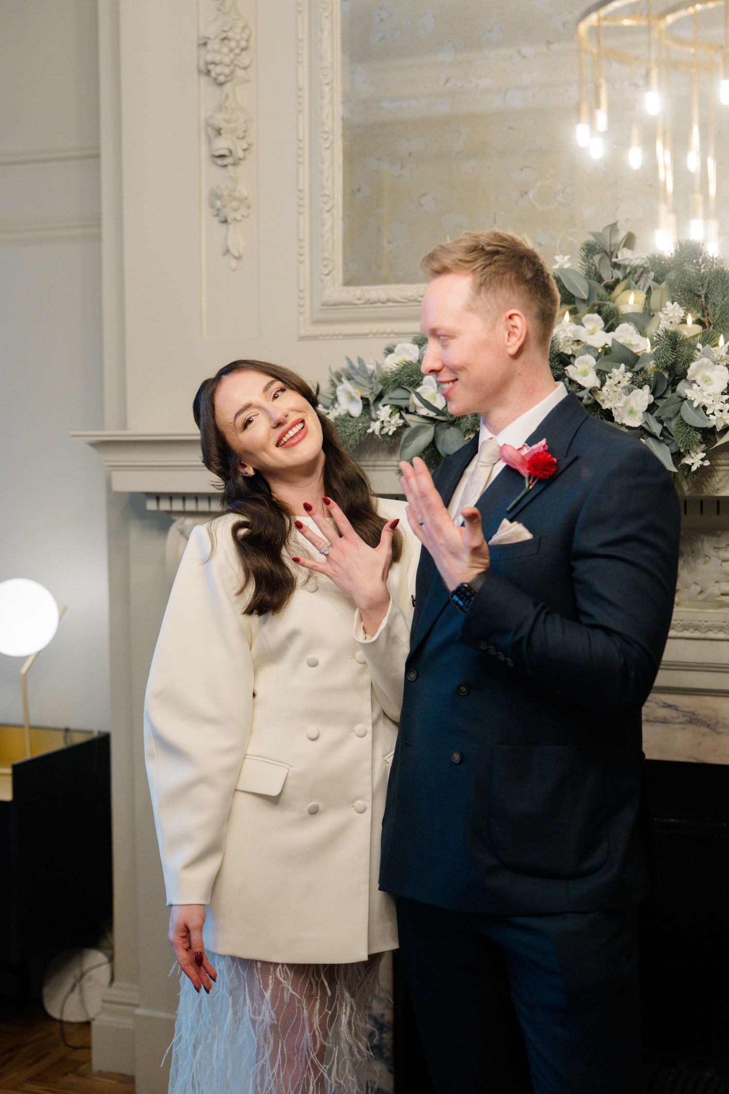 Smiling bride and groom in navy suit show wedding rings by floral fireplace in elegant venue.