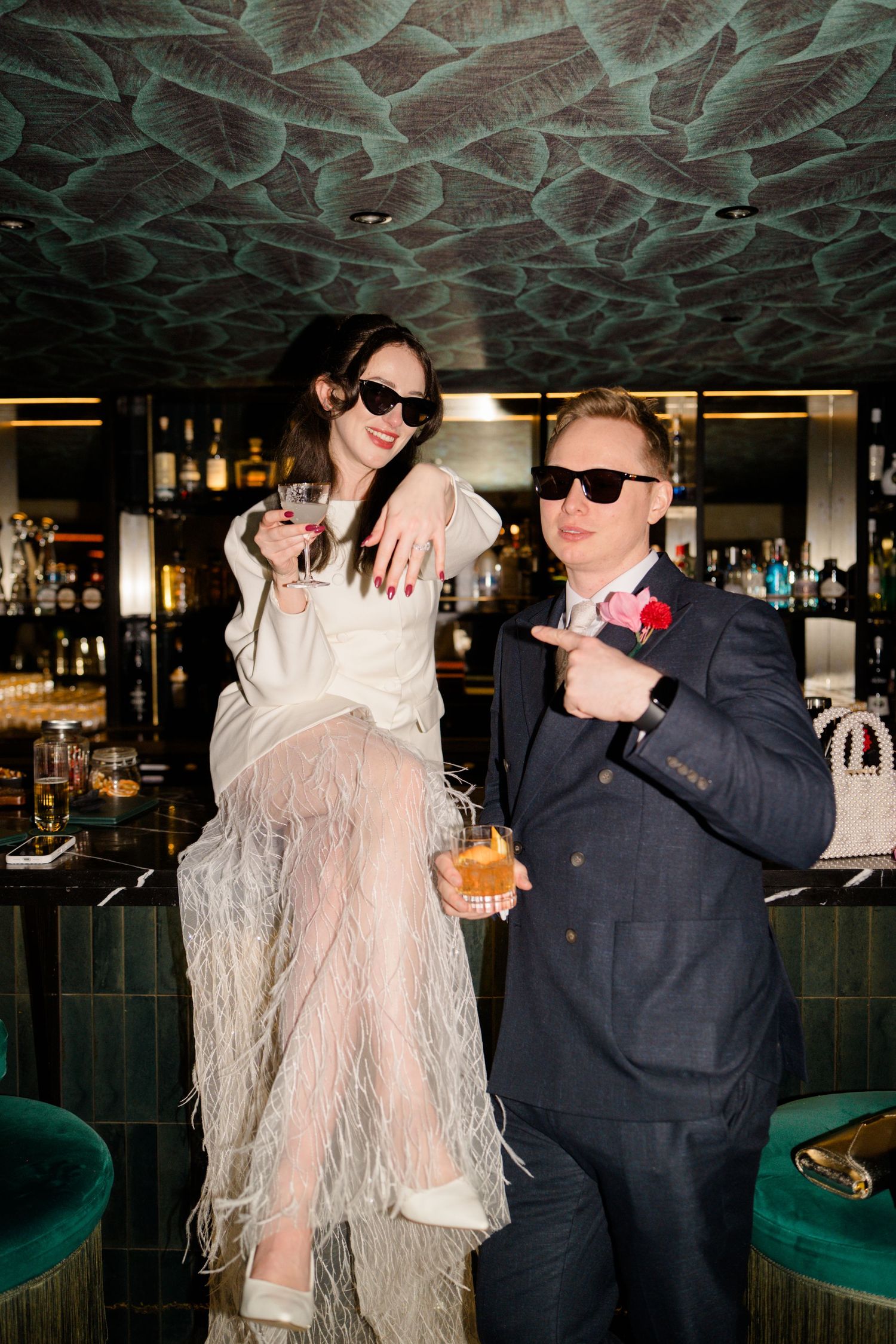 Stylish newlyweds pose at bar wearing sunglasses, bride in feathered skirt holding cocktail with groom.