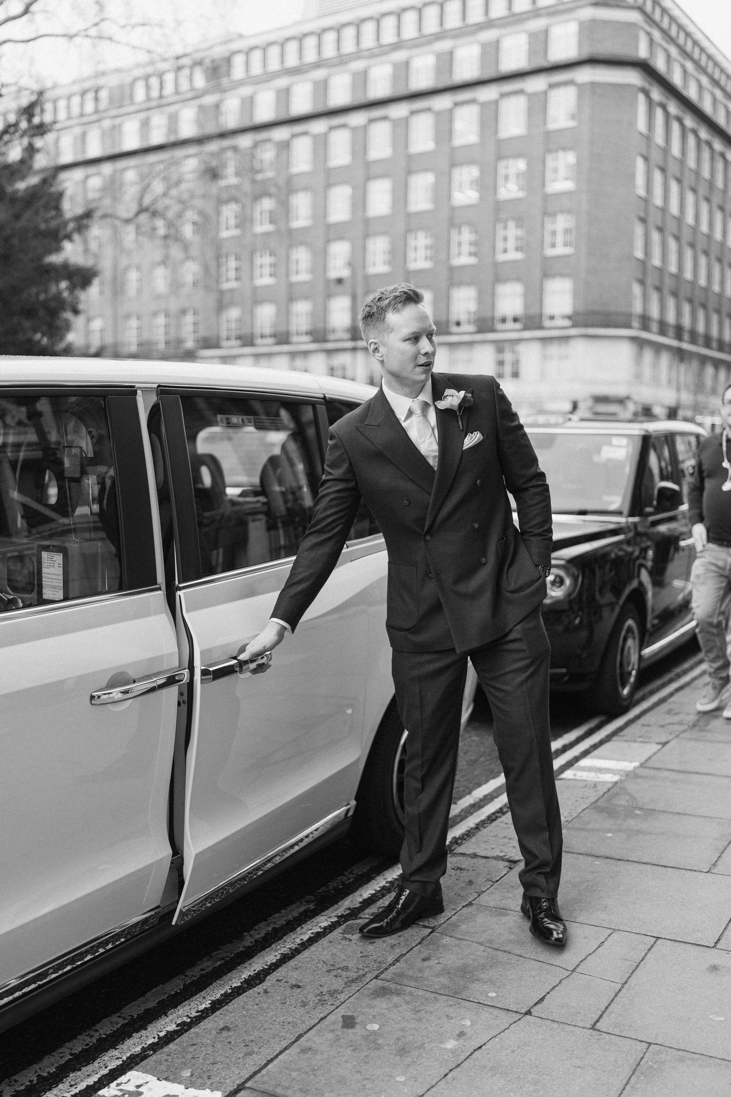 Groom in dark suit opening car door on London street in black and white wedding photo.