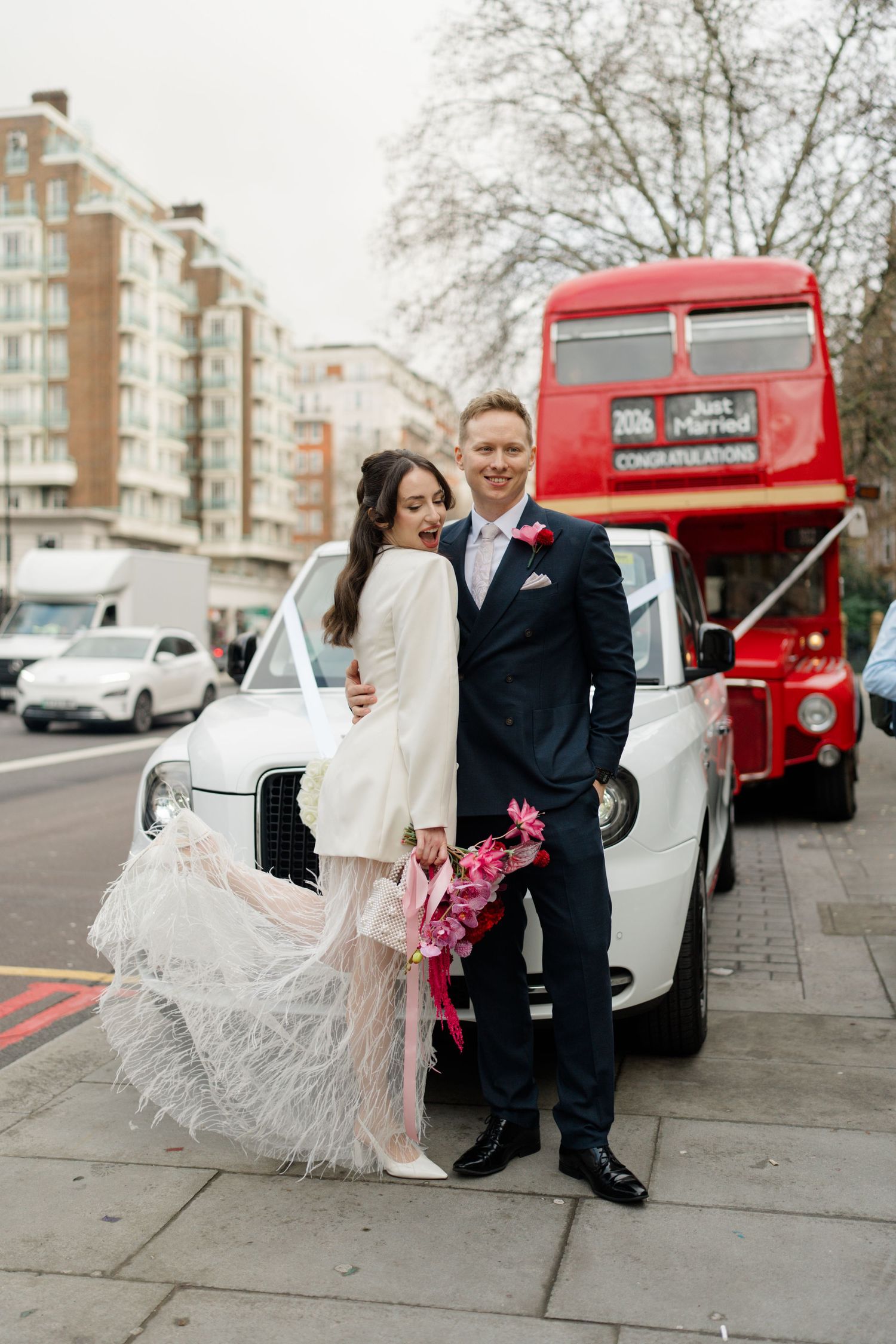 Newlyweds pose in front of a white taxi cab and red double-decker London bus after wedding.