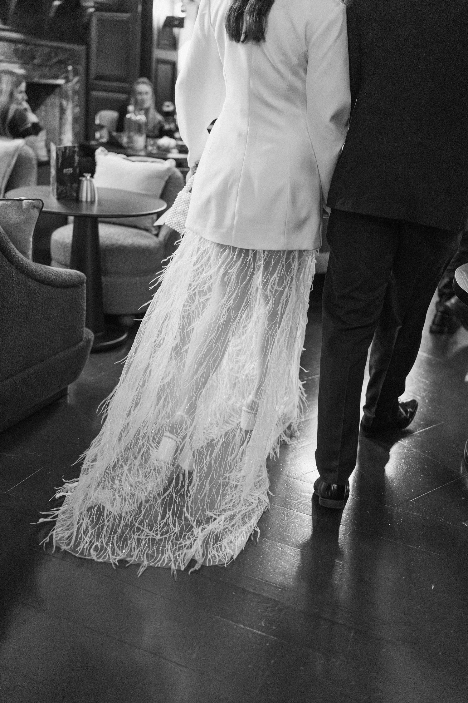 Black and white photo of bride in white jacket and feathered skirt walking with groom in dark suit.