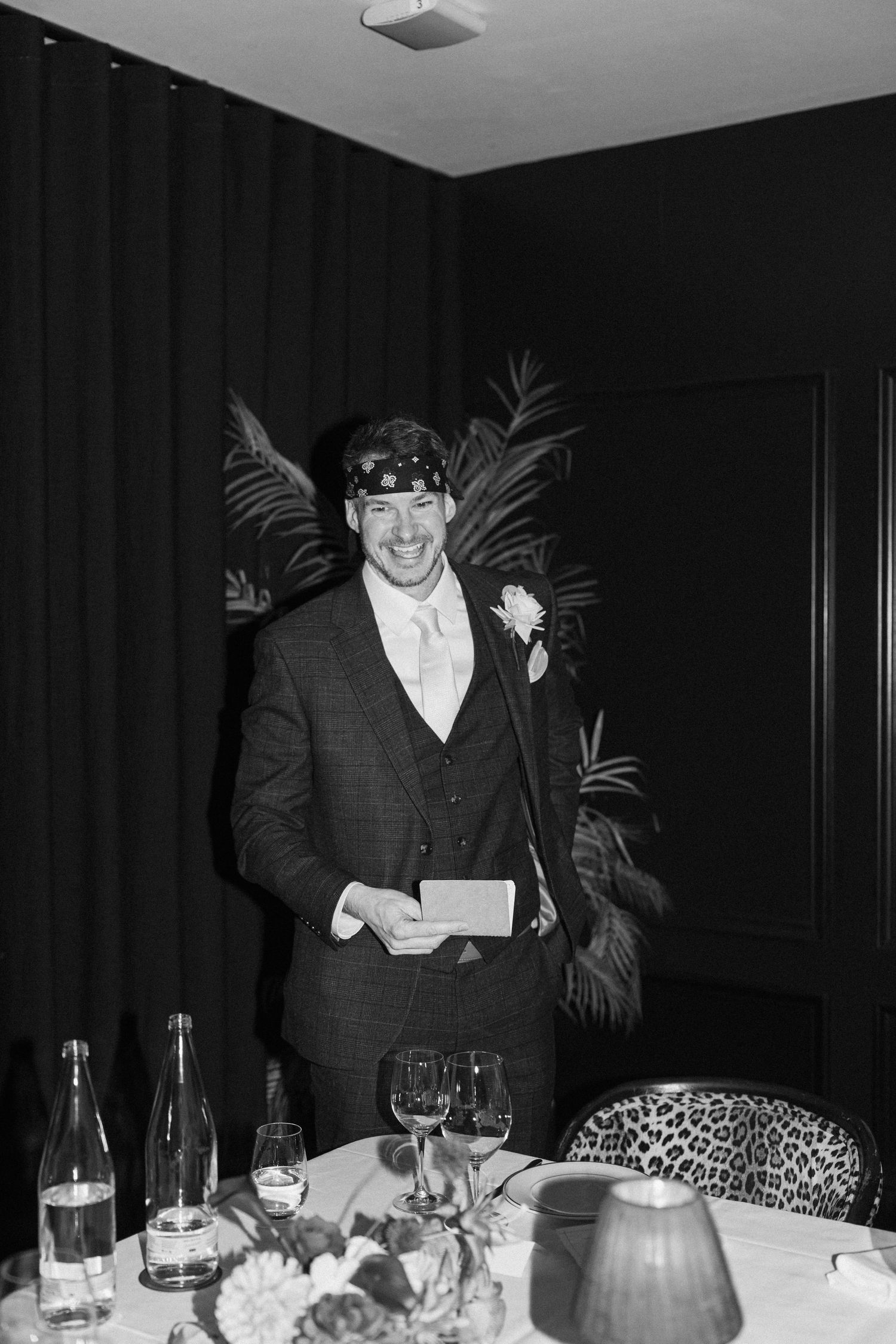 Black and white photo of smiling best man wearing bandana giving speech at elegant wedding reception dinner.