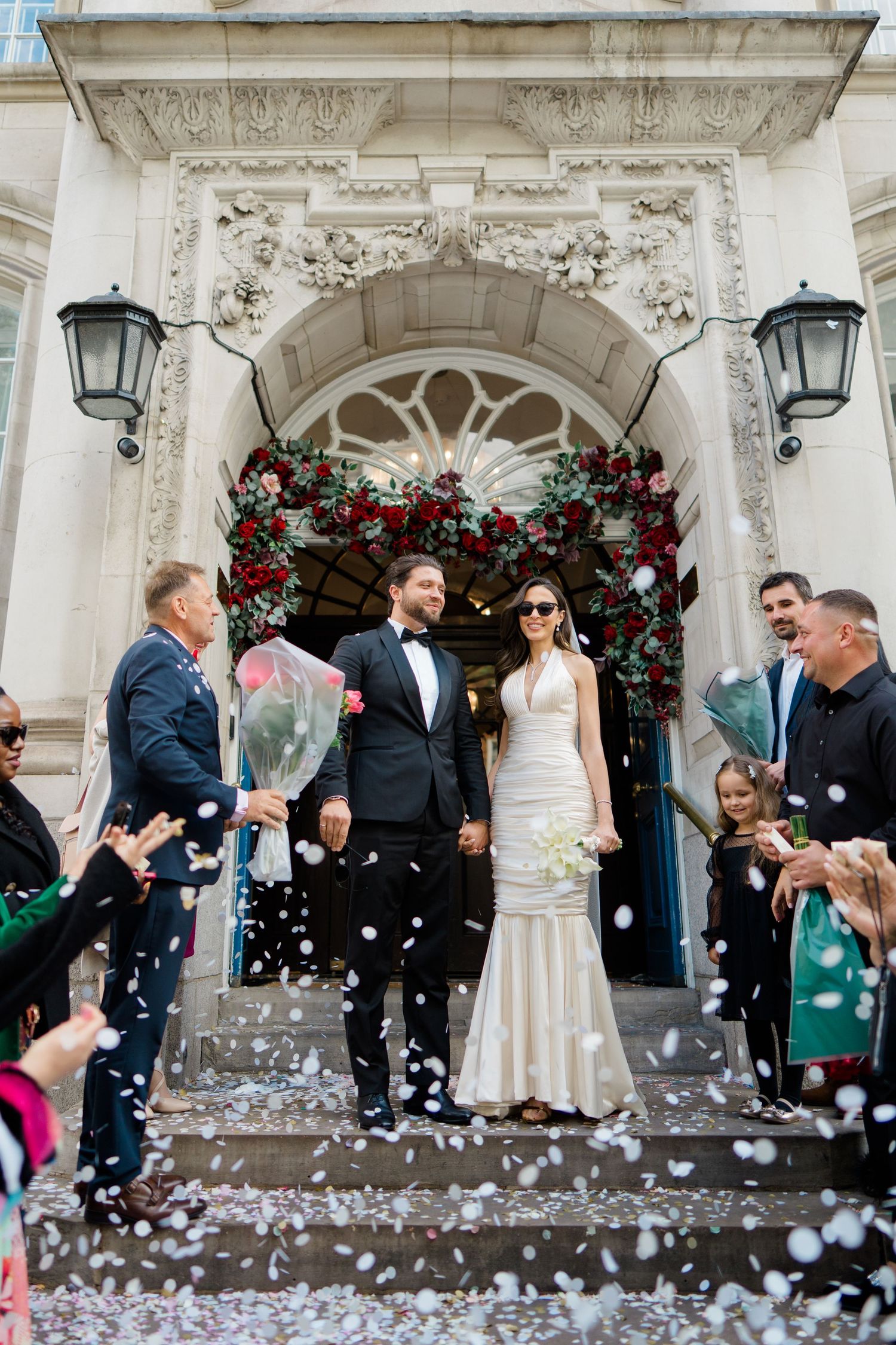 Bride and groom exit ornate building amid confetti shower, guests celebrating their wedding ceremony outdoors.