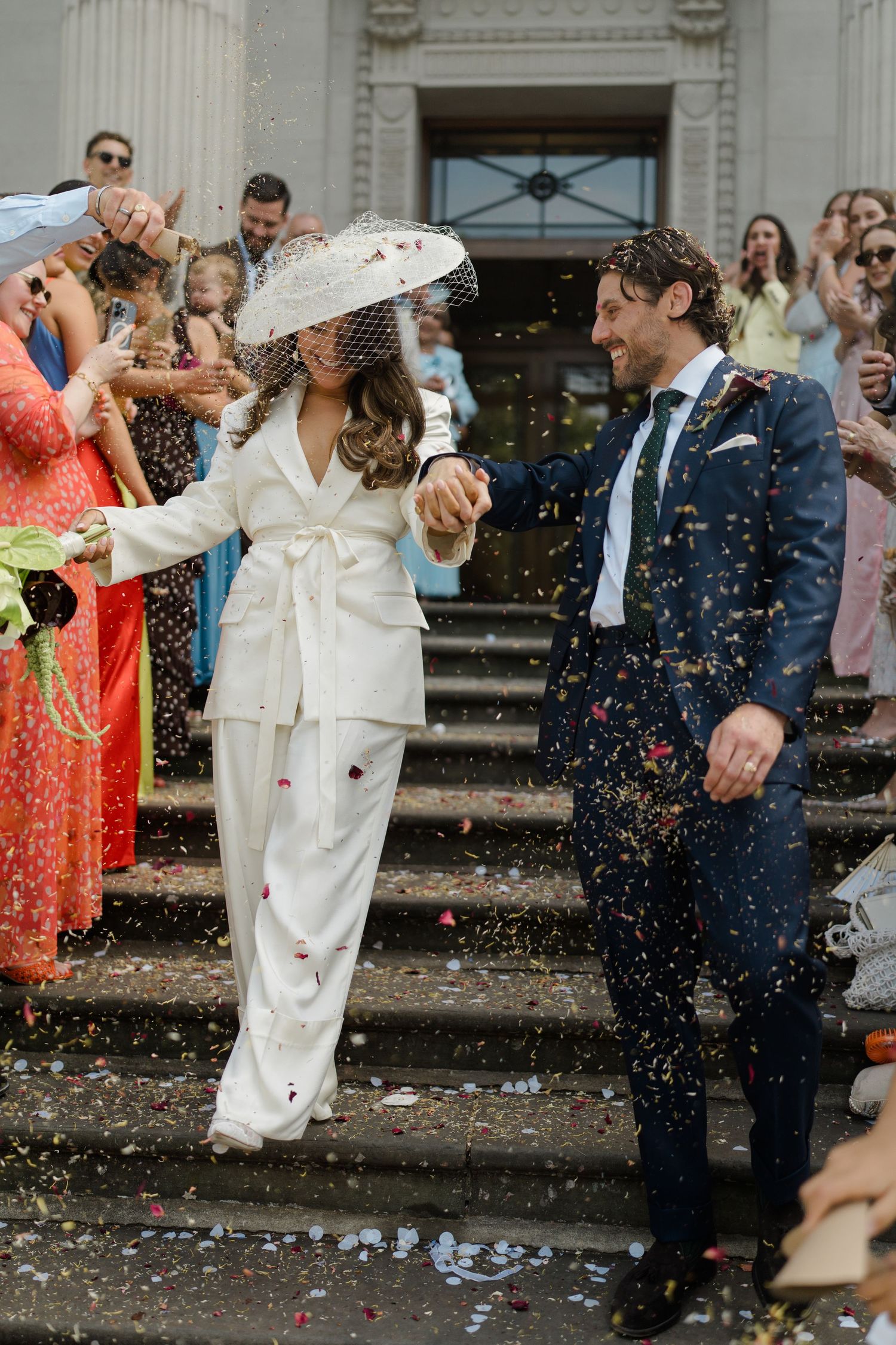 Newlyweds in white suit and navy tuxedo exit venue steps as guests throw colorful confetti.