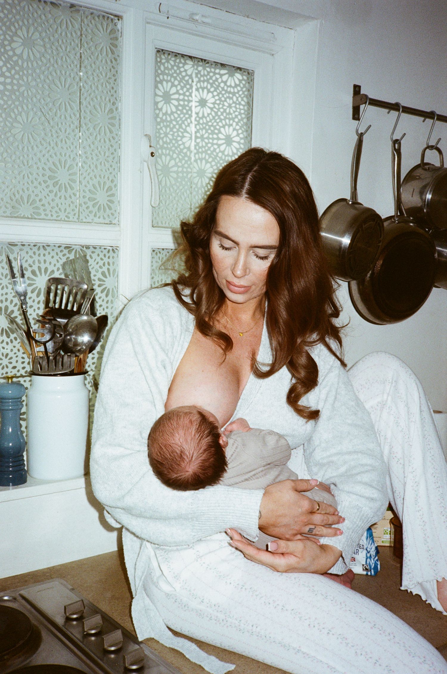 A woman in white robe breastfeeding her newborn baby while sitting in a cozy kitchen setting.