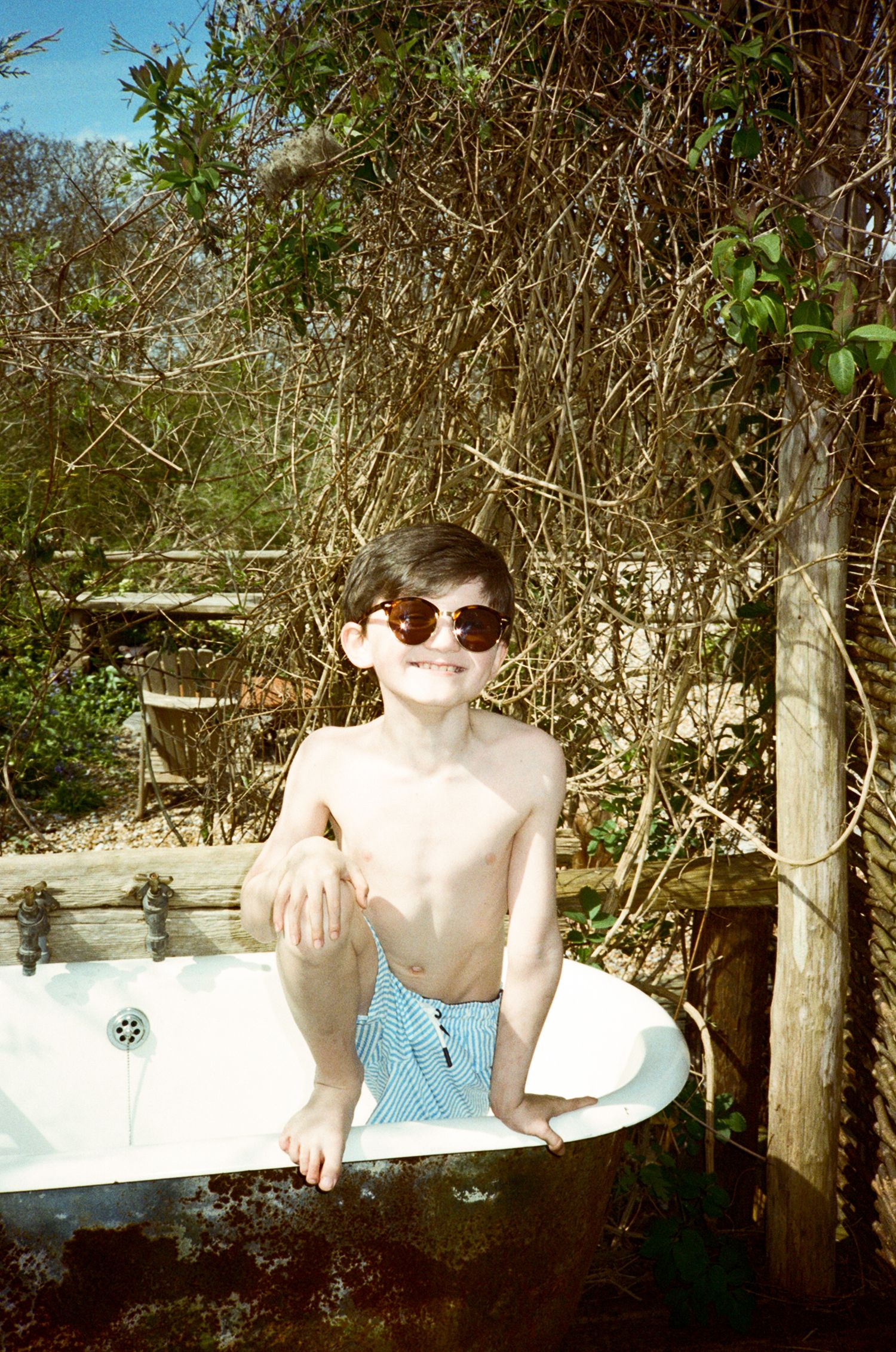 A young boy wearing sunglasses and swim trunks sits in an outdoor bathtub surrounded by lush garden greenery.