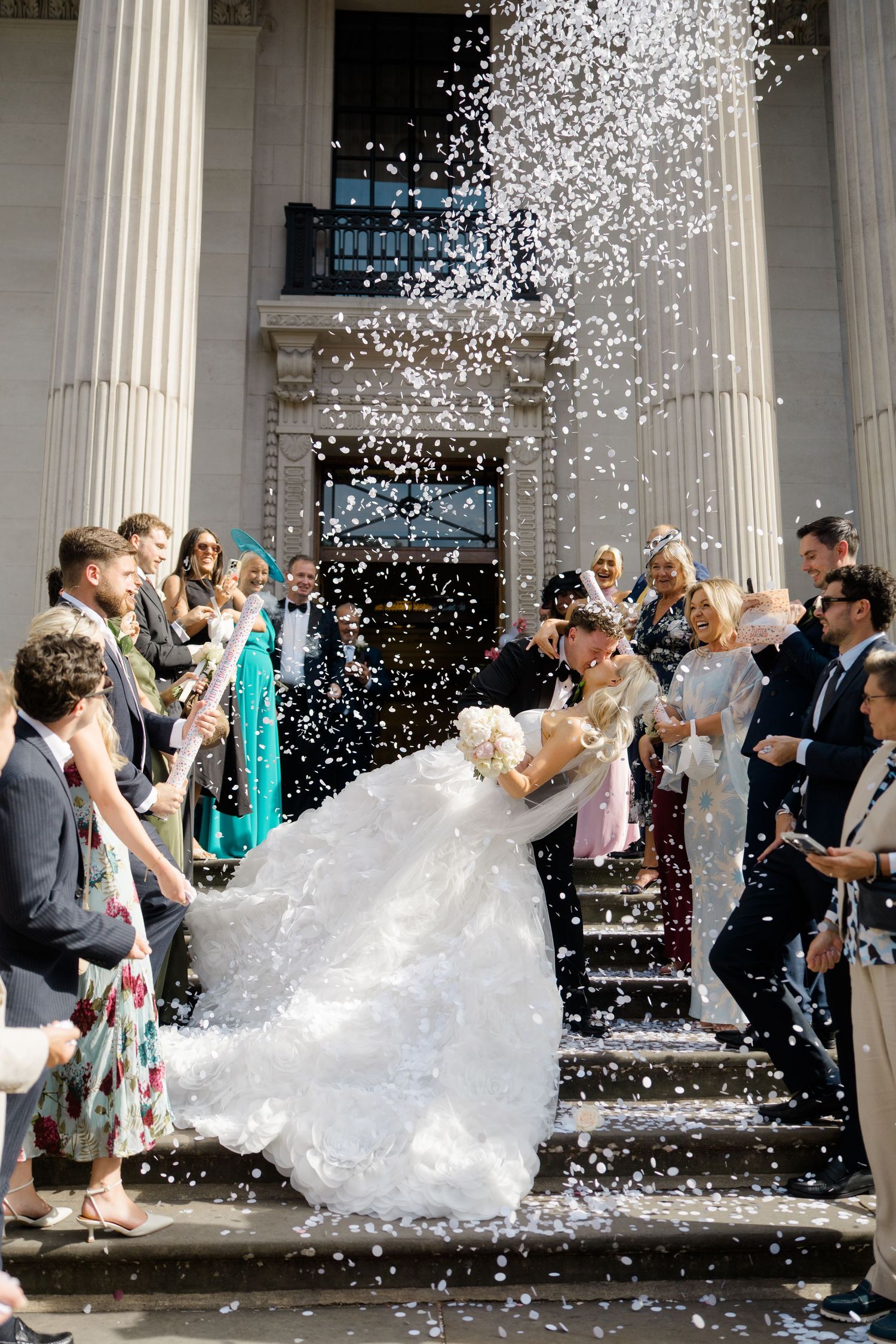 Bride and groom share a kiss on church steps as confetti showers over them surrounded by wedding guests.