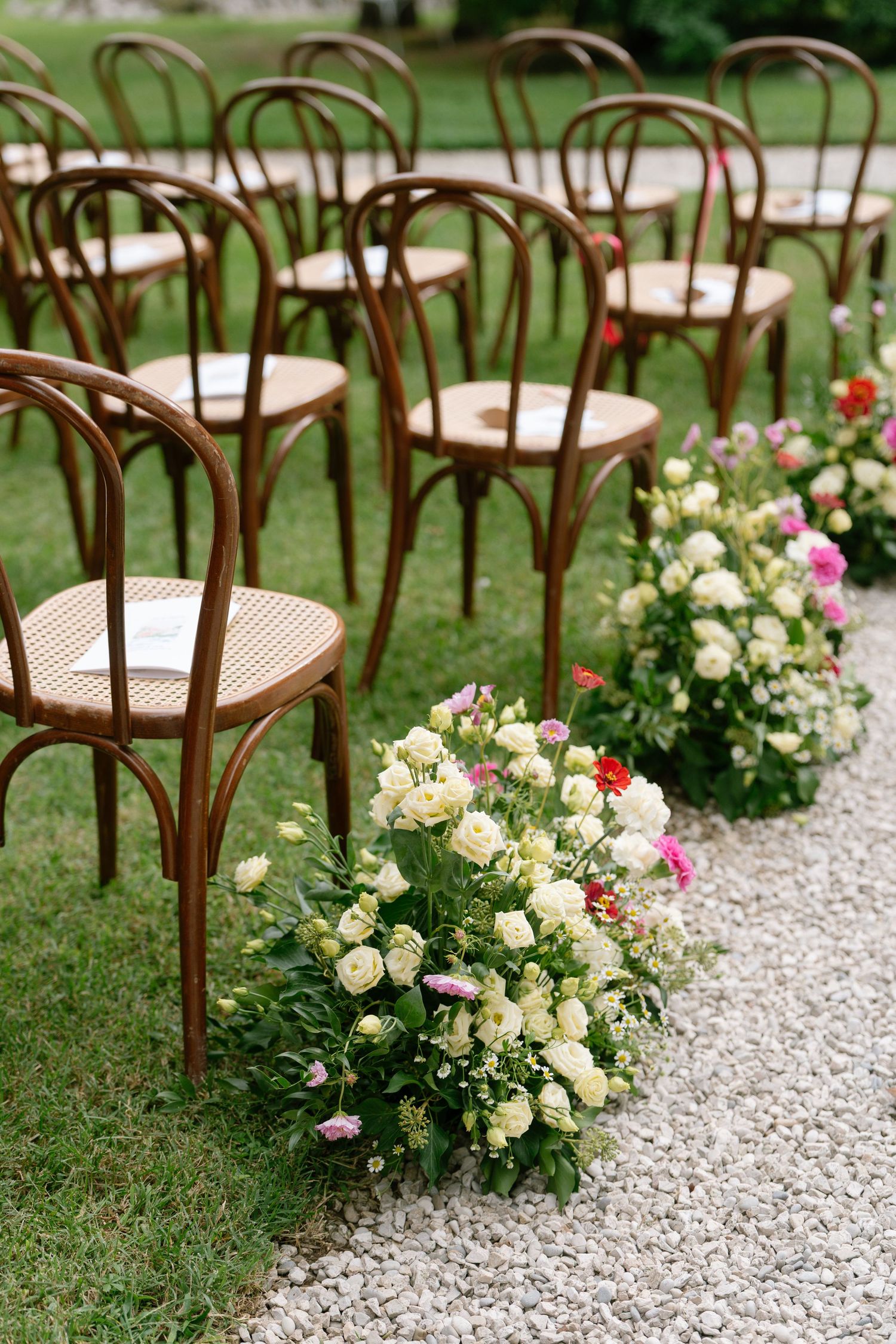 Bentwood chairs line a gravel aisle with fresh floral arrangements for an outdoor wedding.