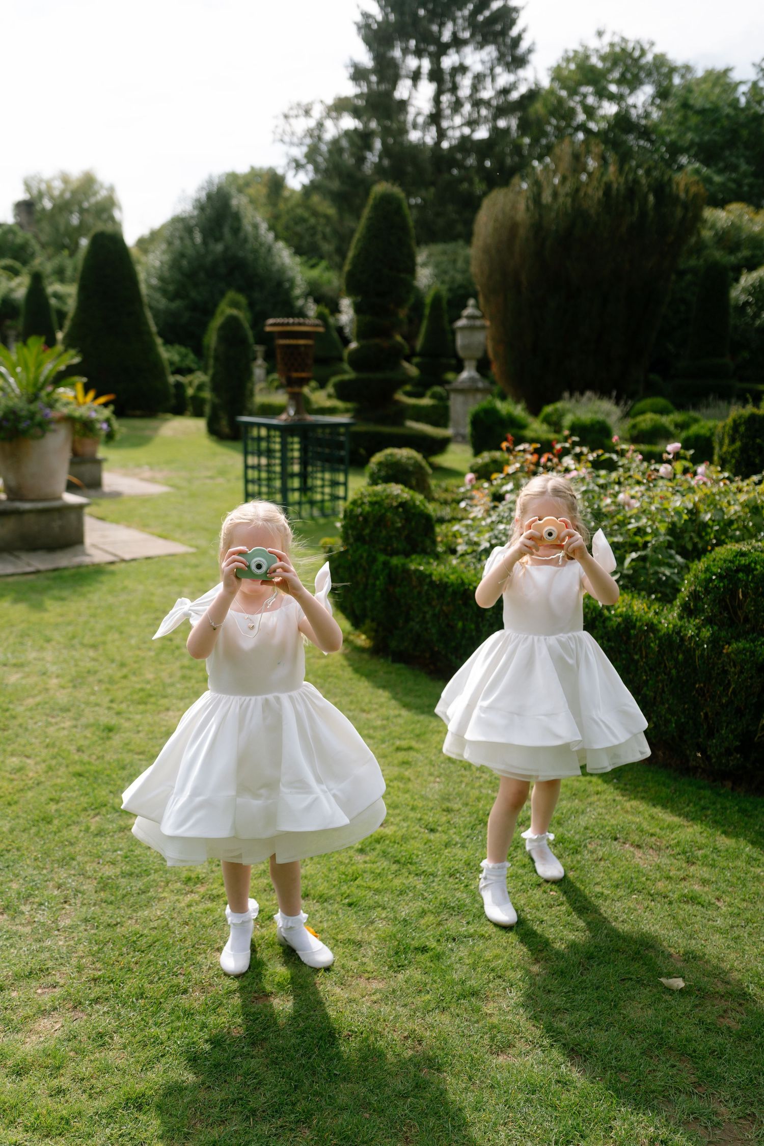 Little ones in white dresses play on a manicured garden lawn with topiary.