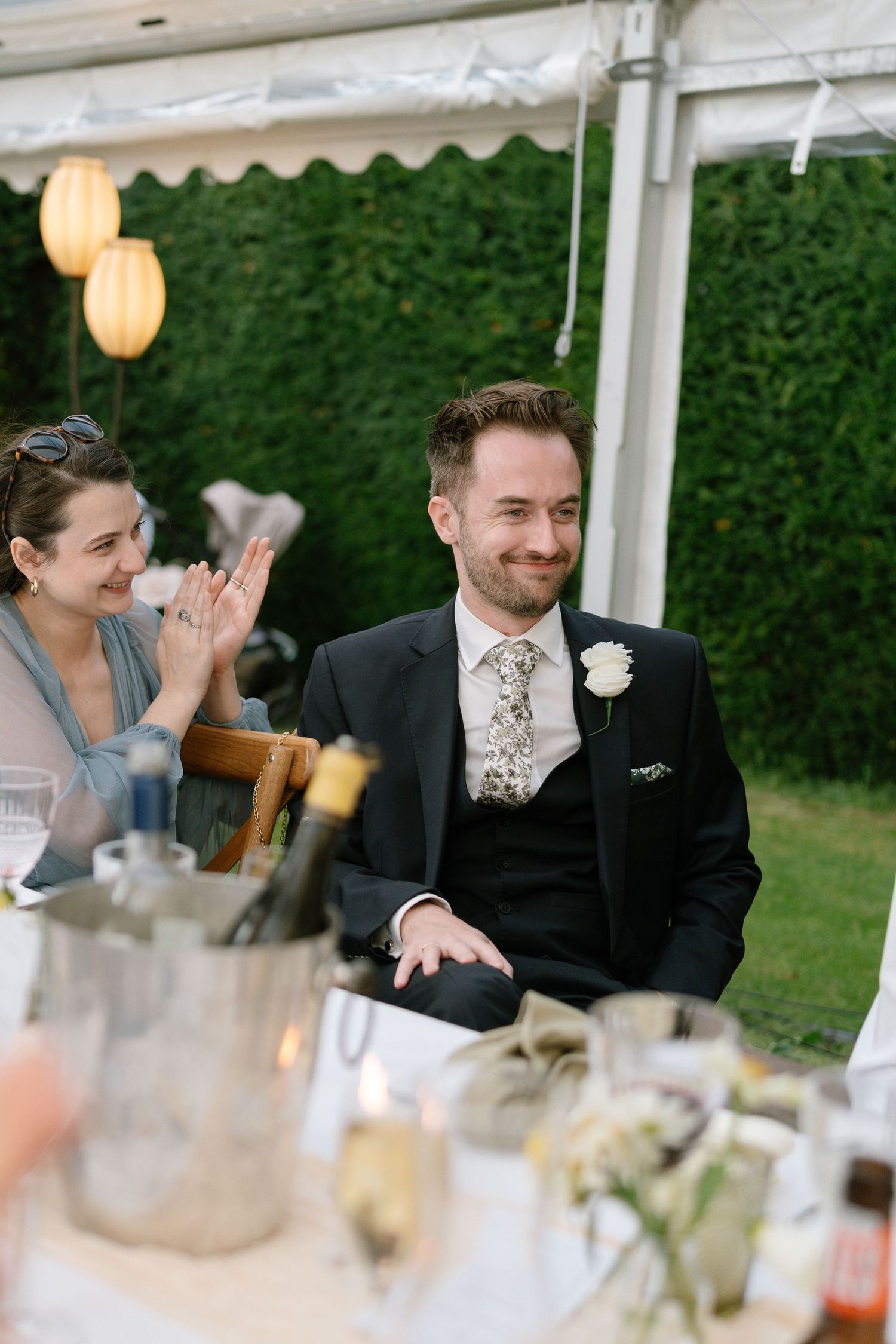 Wedding guest smiling during reception under tent with green hedge backdrop and paper lanterns.