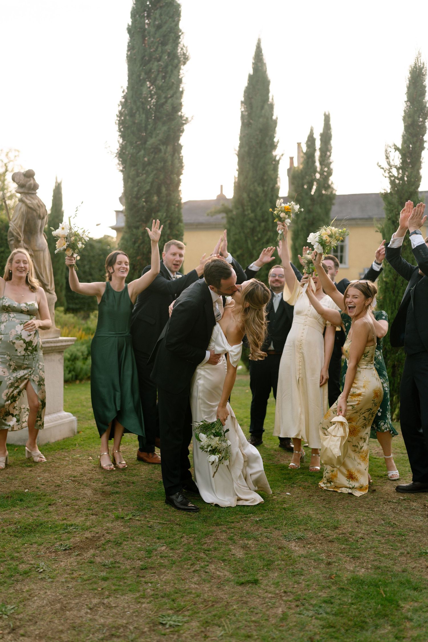 Wedding group raises glasses in celebratory toast on lawn with cypress trees.