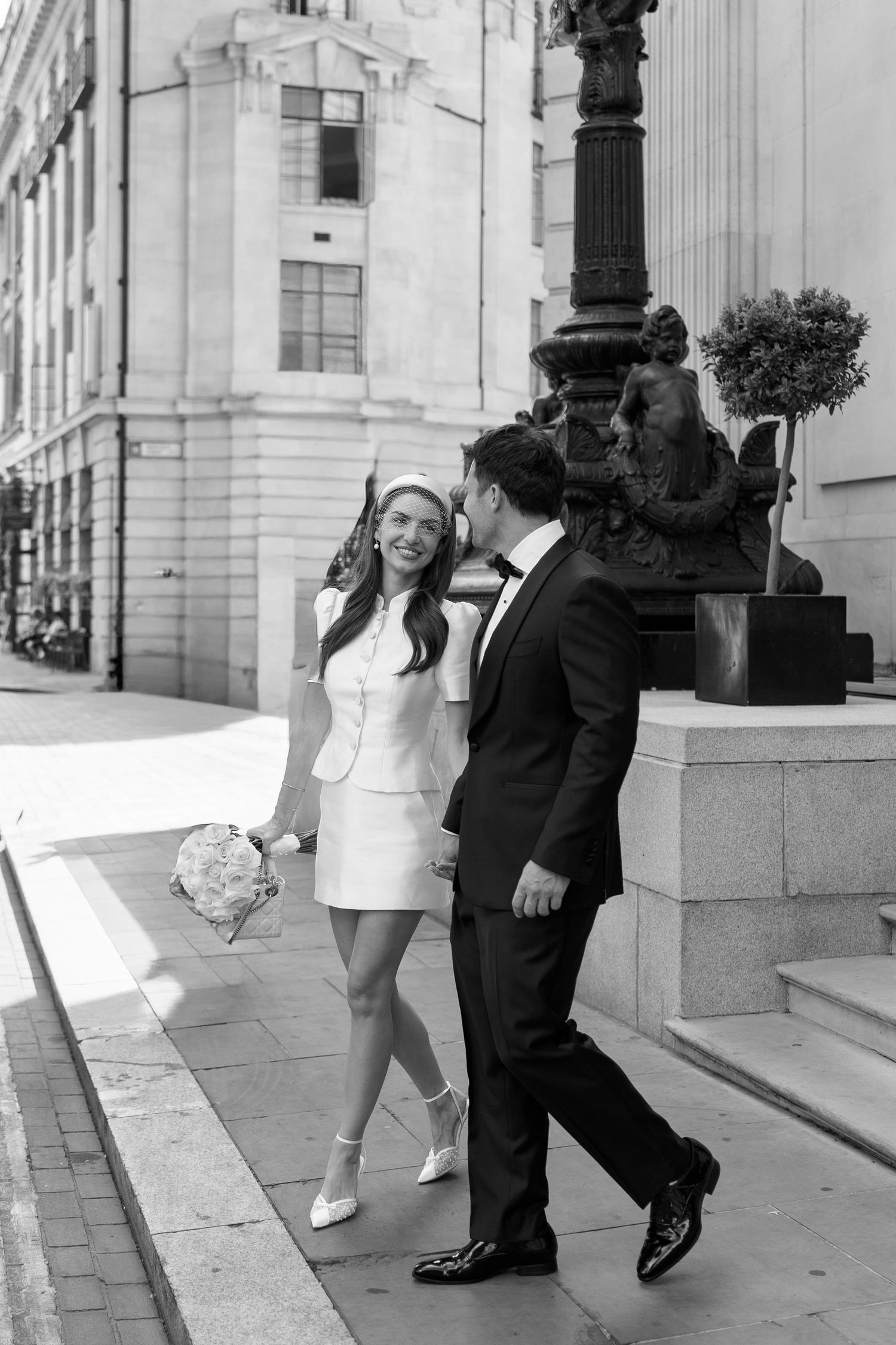 Black and white photo of a smiling bride in mini dress holding bouquet walking with groom in tuxedo on city sidewalk.