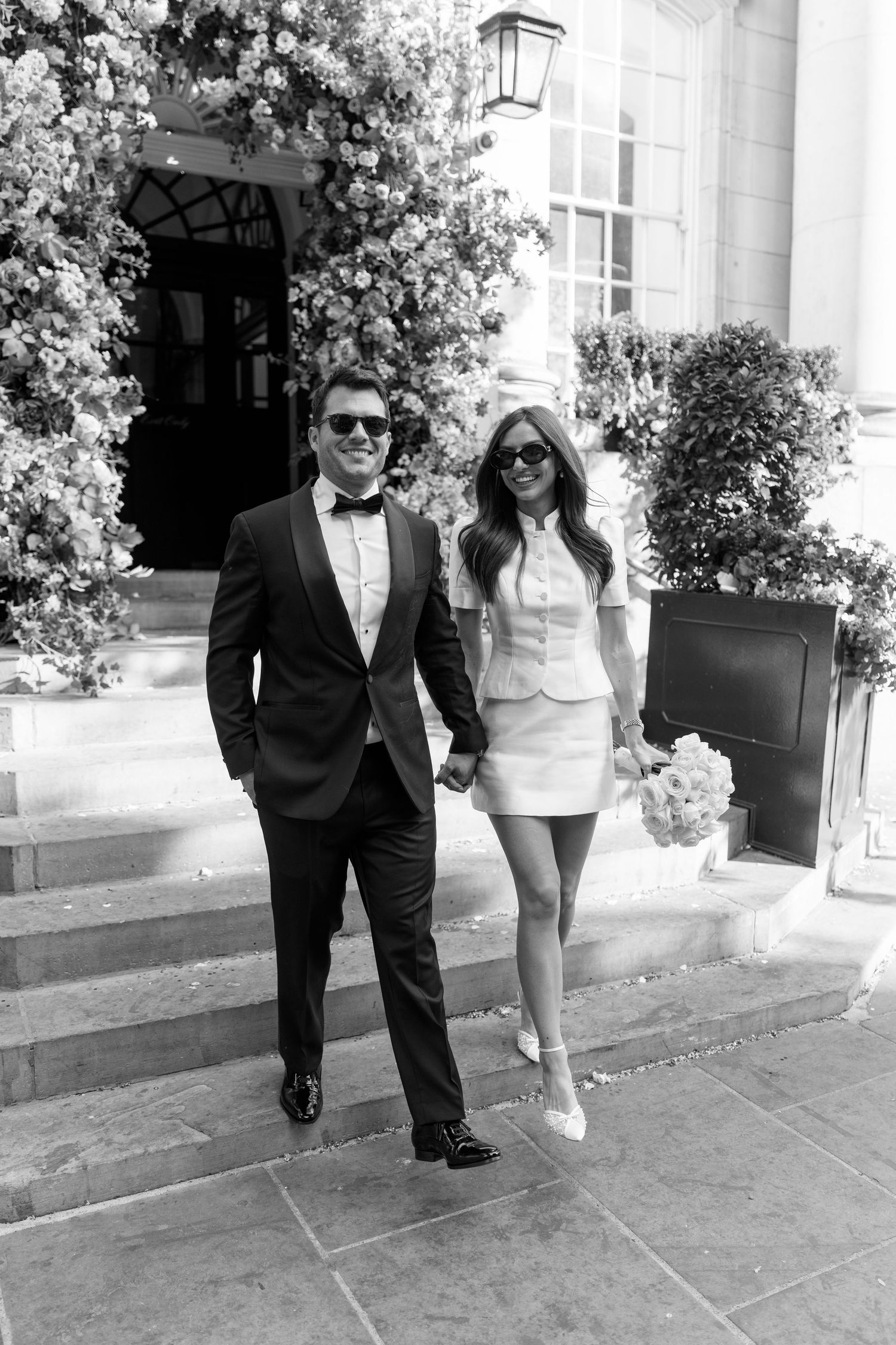 Black and white photo of an elegant couple in formal attire holding hands on steps outside a floral-decorated building.
