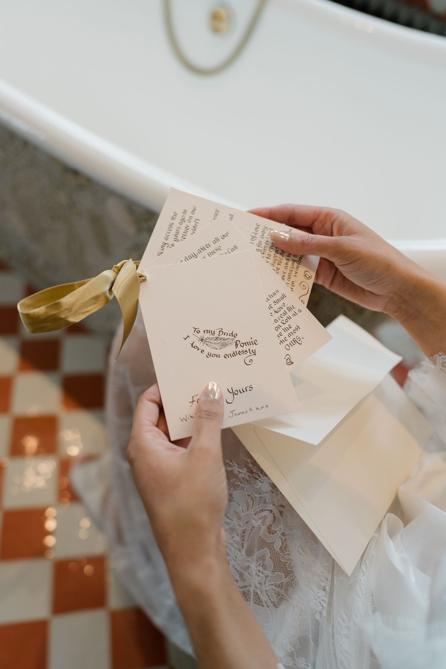 Bride in lace robe reading handwritten wedding vow cards beside a freestanding bathtub.