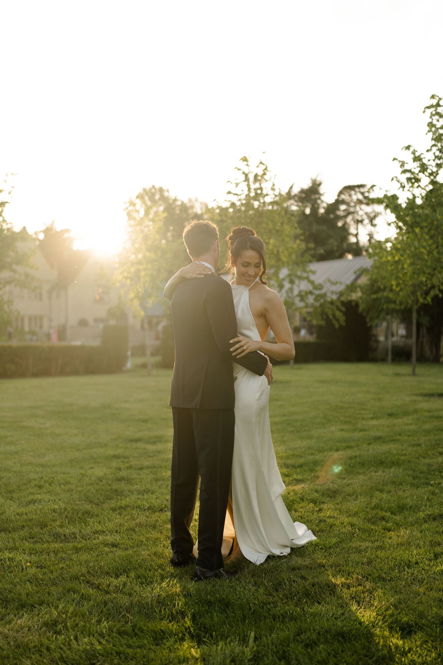 Bride and groom embrace on lawn during golden sunset hour at elegant outdoor wedding venue.