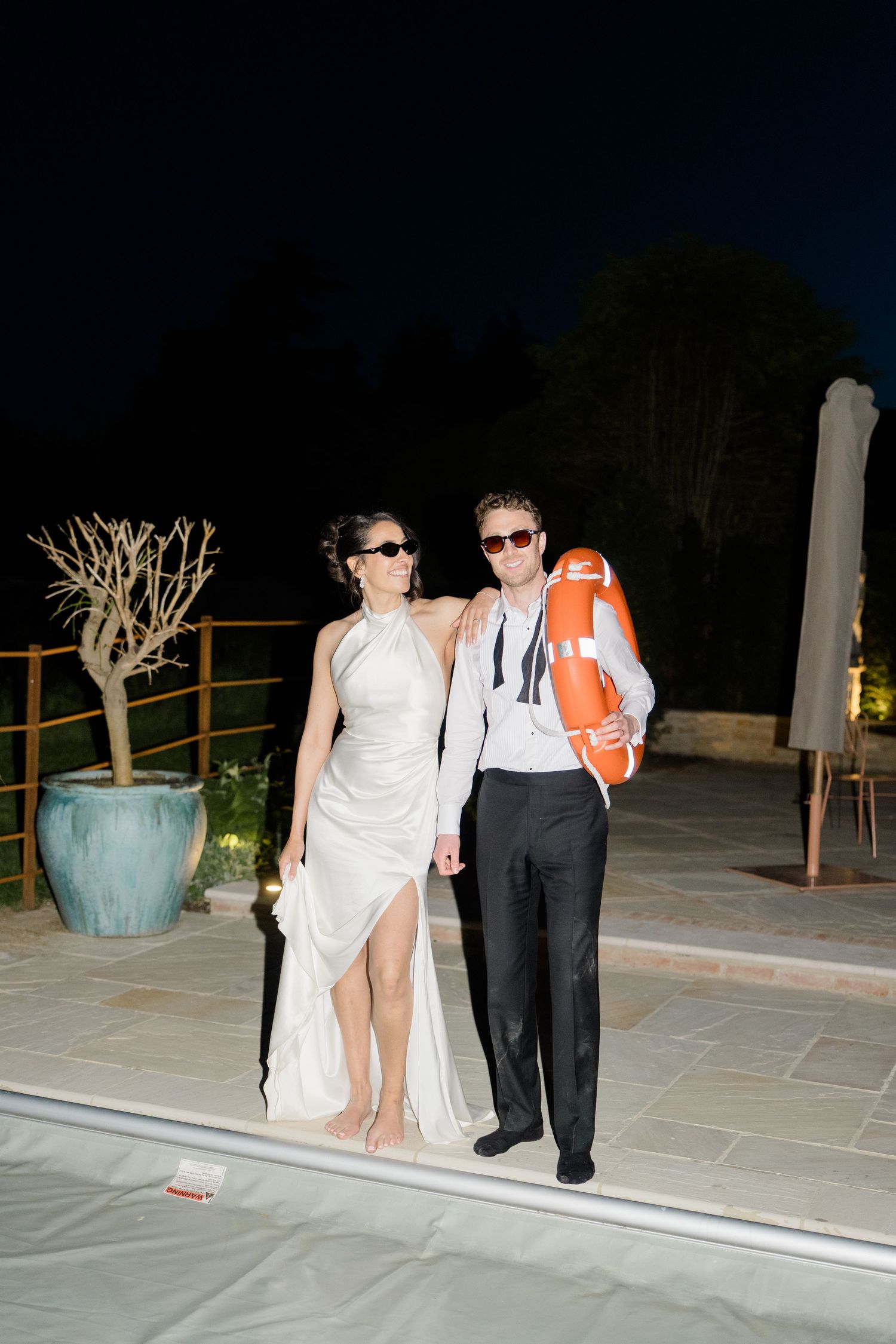 Bride and groom pose poolside at night wearing sunglasses, groom holding a life preserver.