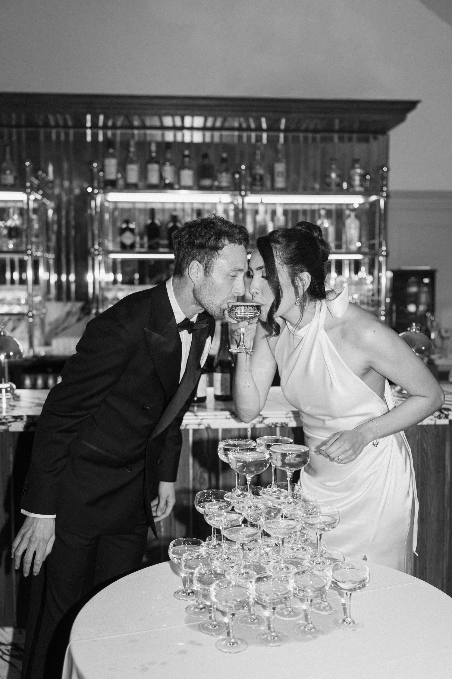 Black and white photo of bride and groom sharing champagne at a coupe tower at the bar.