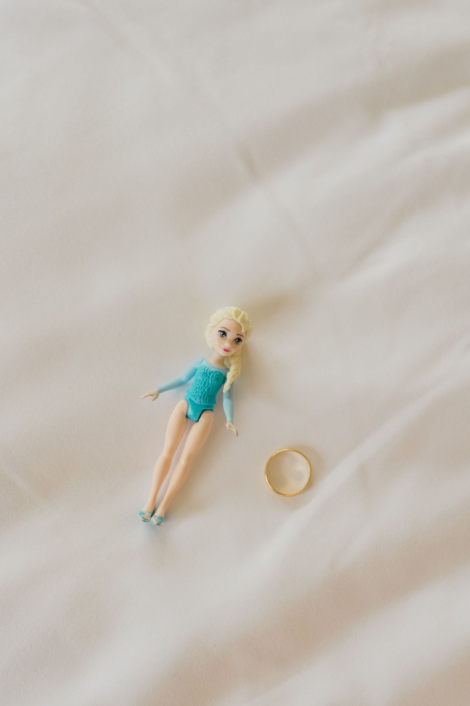 Elsa Disney princess doll lying next to a gold wedding ring on white fabric bedding.
