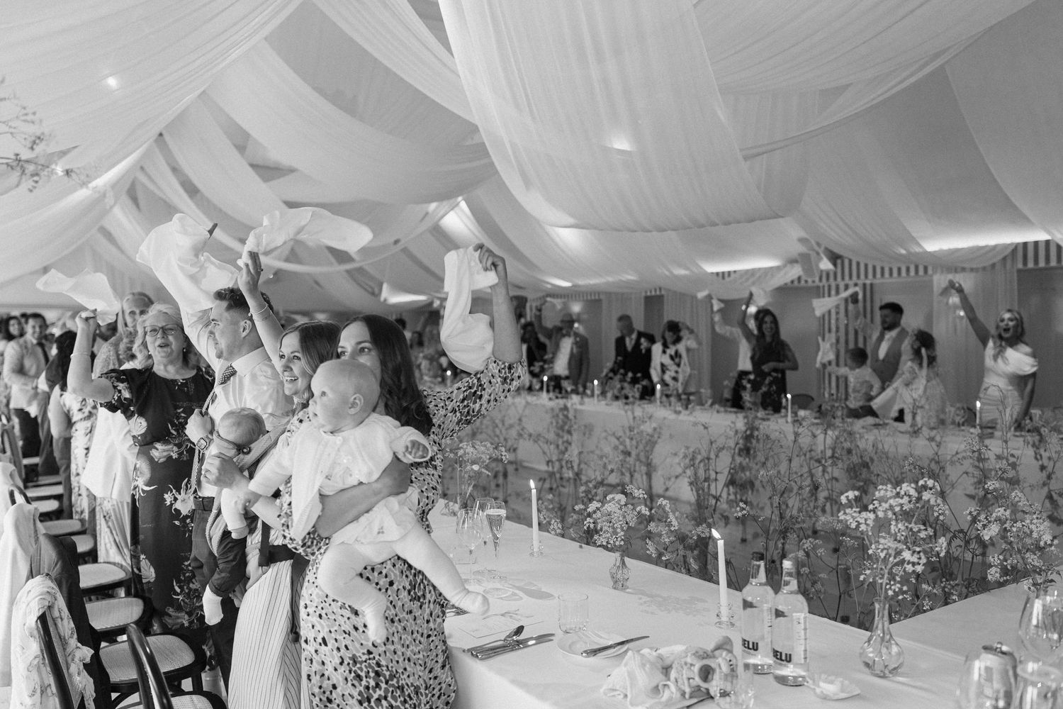 Black and white photo of wedding guests celebrating under draped marquee ceiling at lively reception.