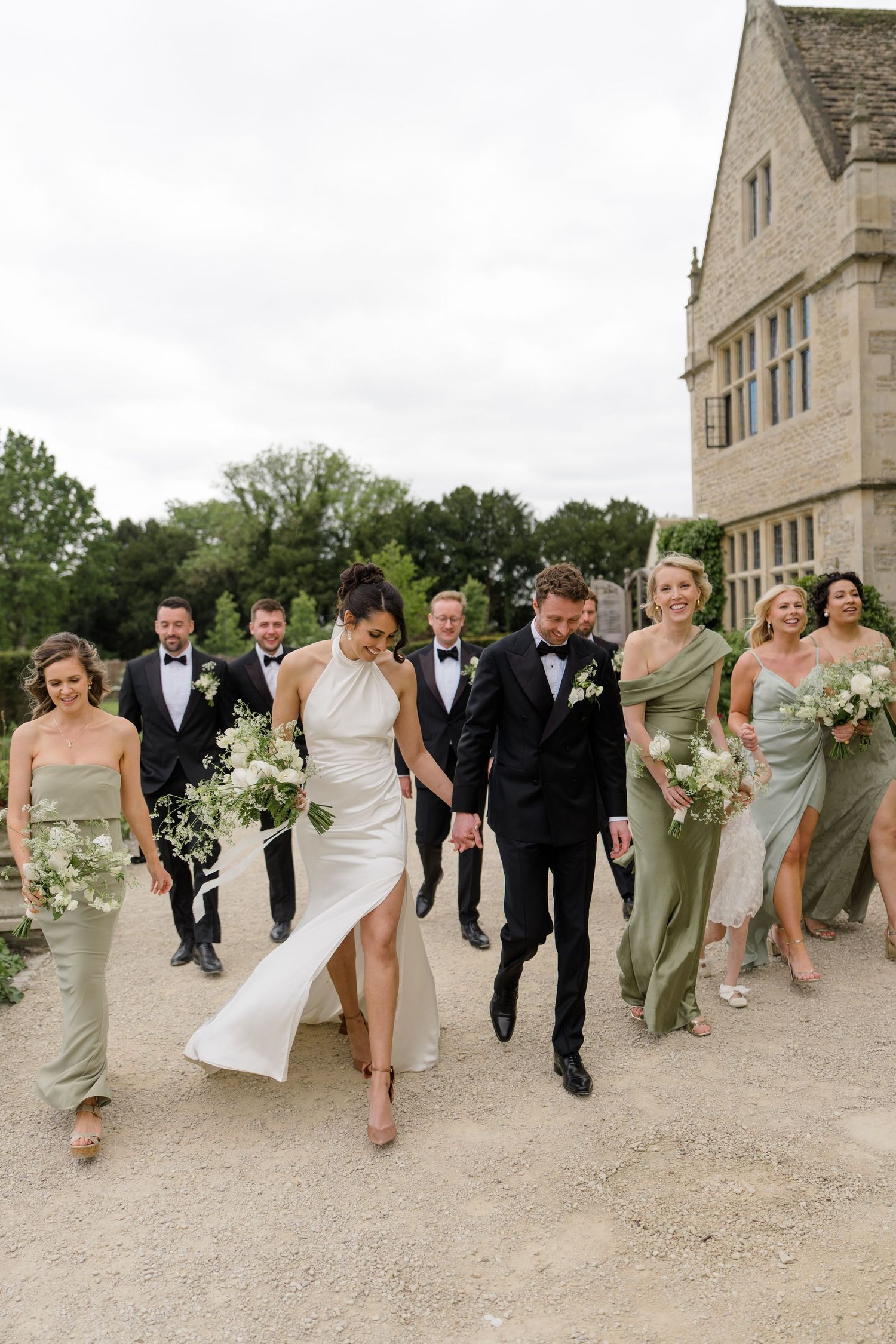 Wedding party walks together outside historic stone manor, bridesmaids in sage green dresses.
