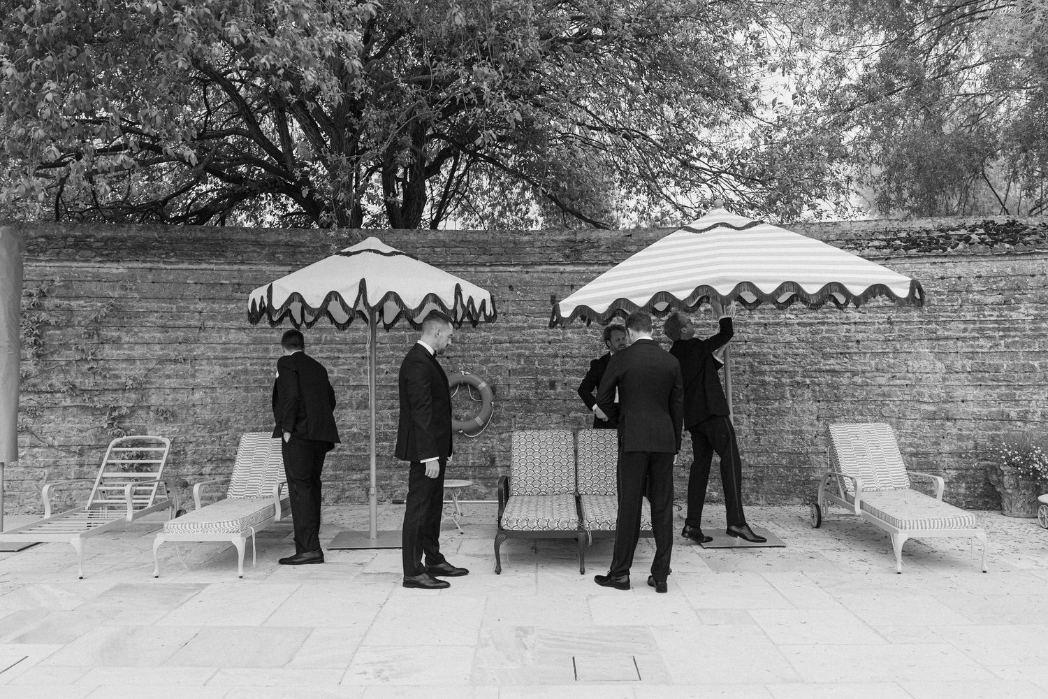 Black and white photo of groomsmen in suits gathered by striped umbrellas at pool area.