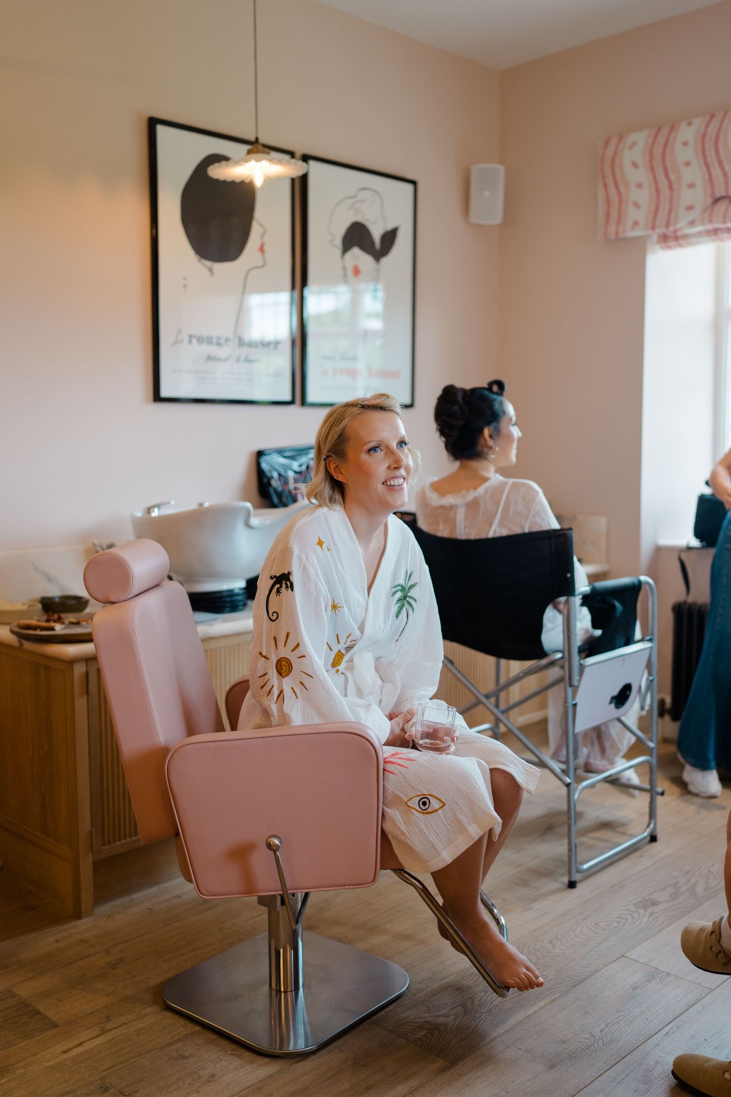 Bride in white robe smiling in pink salon chair during wedding morning preparations.