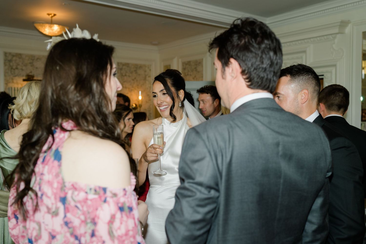Smiling bride in white gown mingles with wedding guests during indoor reception cocktail hour.