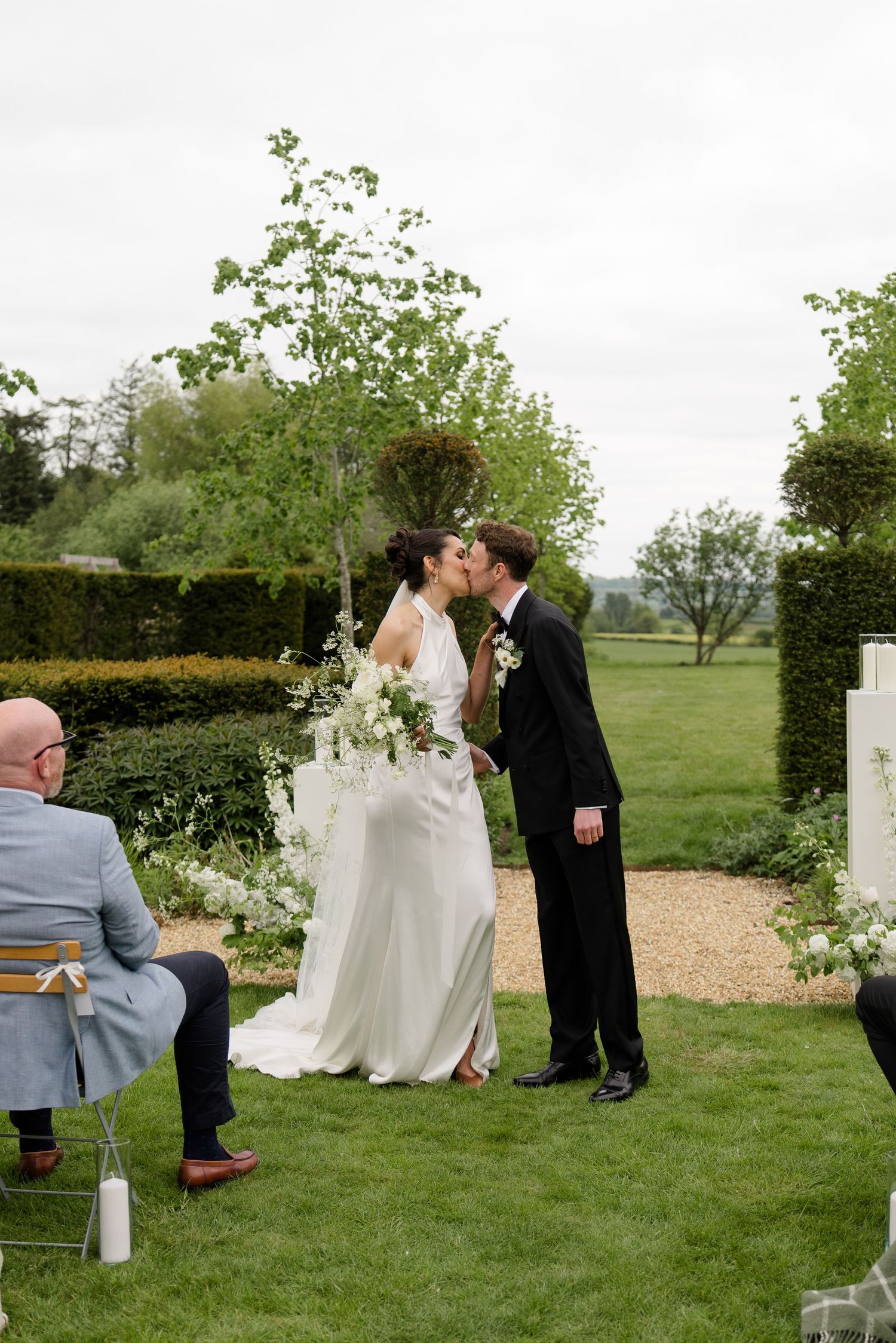 Bride and groom share first kiss outdoors in lush garden setting during wedding ceremony.