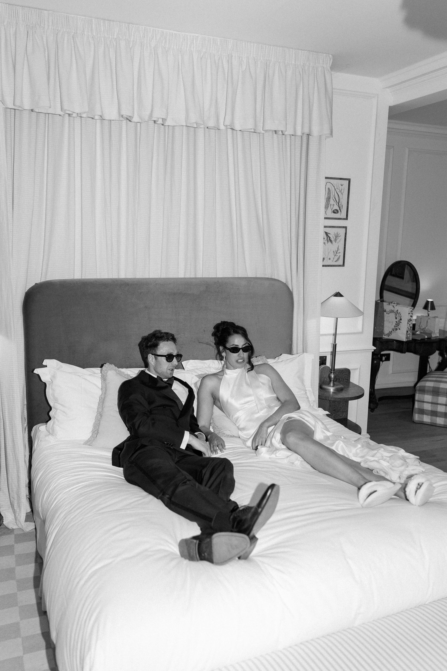 Black and white photo of bride and groom relaxing on hotel bed wearing sunglasses after wedding celebration.