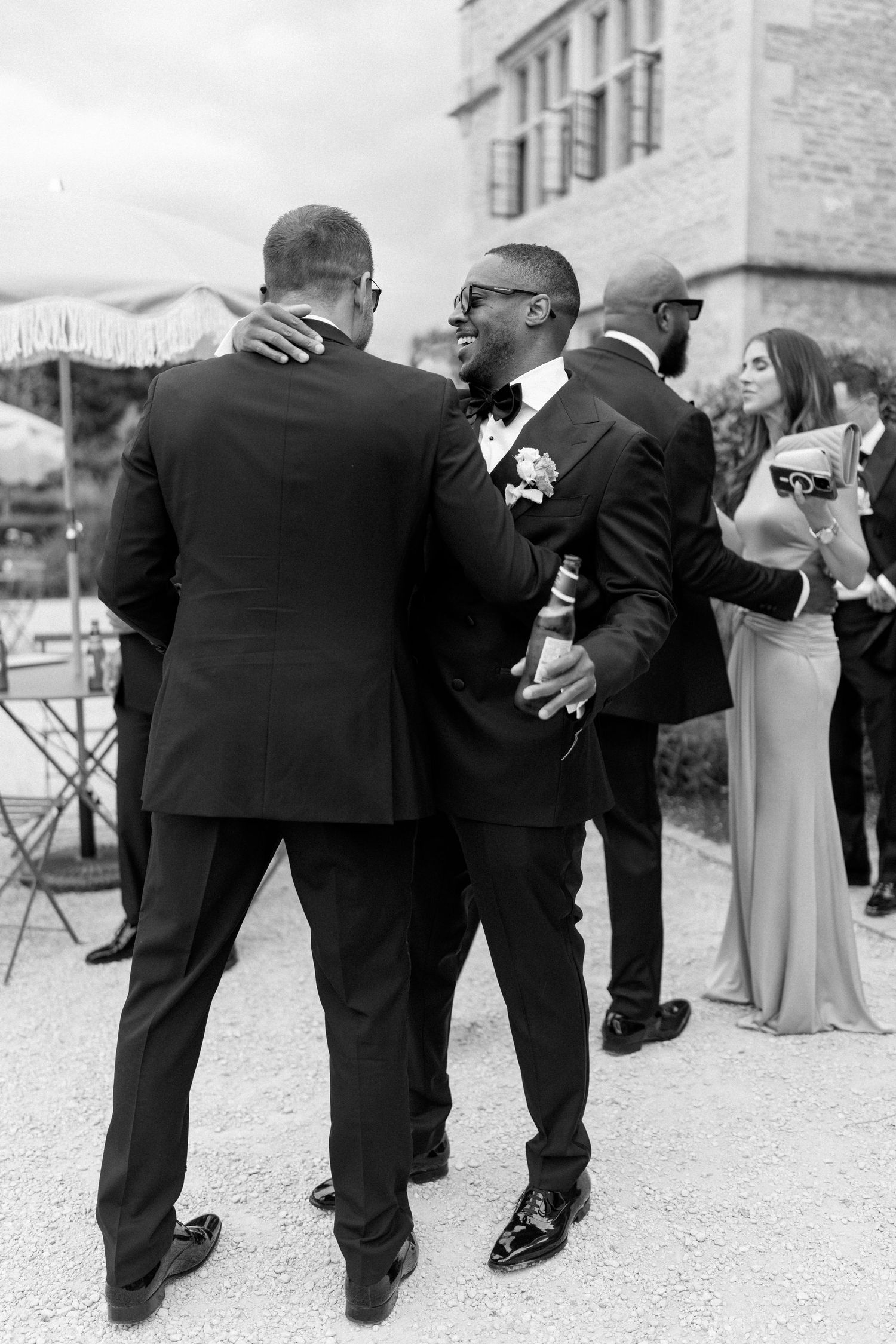 Two men in tuxedos embracing at an outdoor wedding reception in black and white.