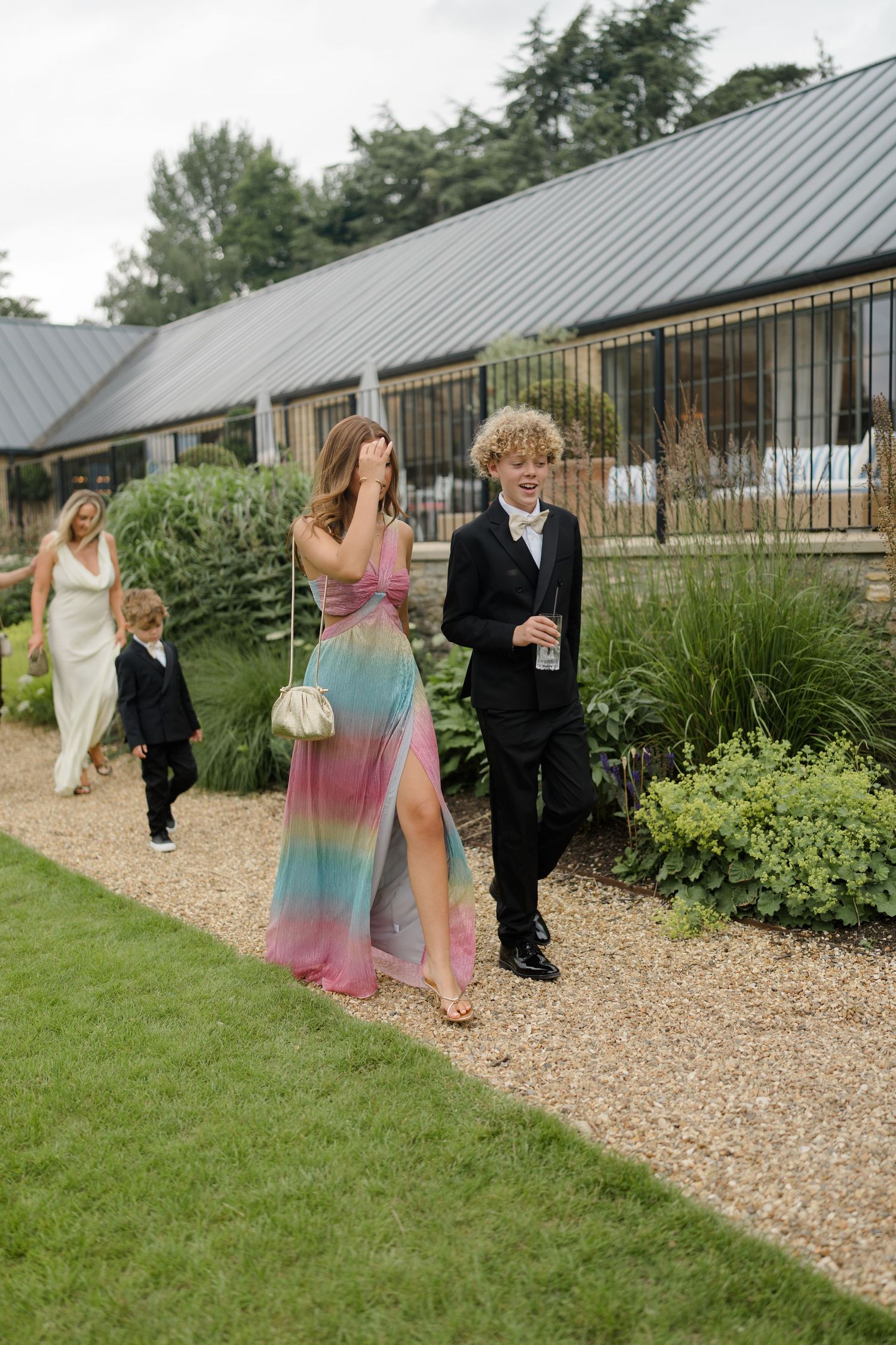 Wedding guests walking on gravel path, woman in colorful rainbow gown with young man in suit.