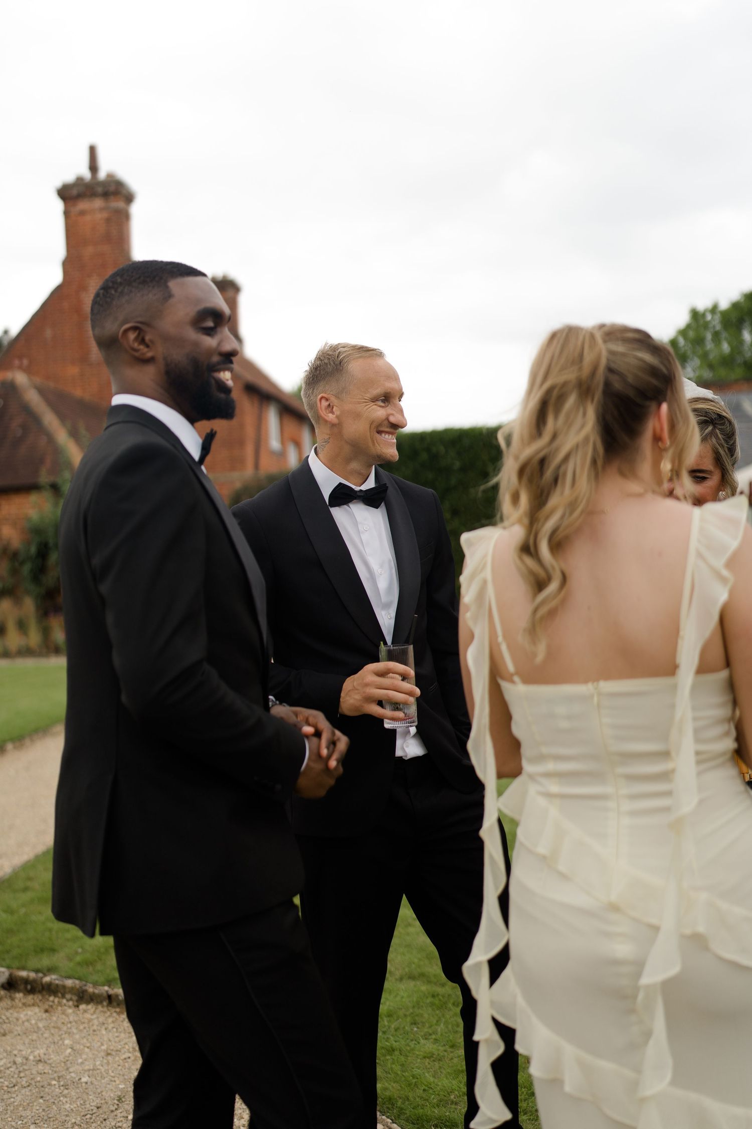 Two smiling groomsmen in black tuxedos chat with the bride in a ruffled white wedding dress outdoors.