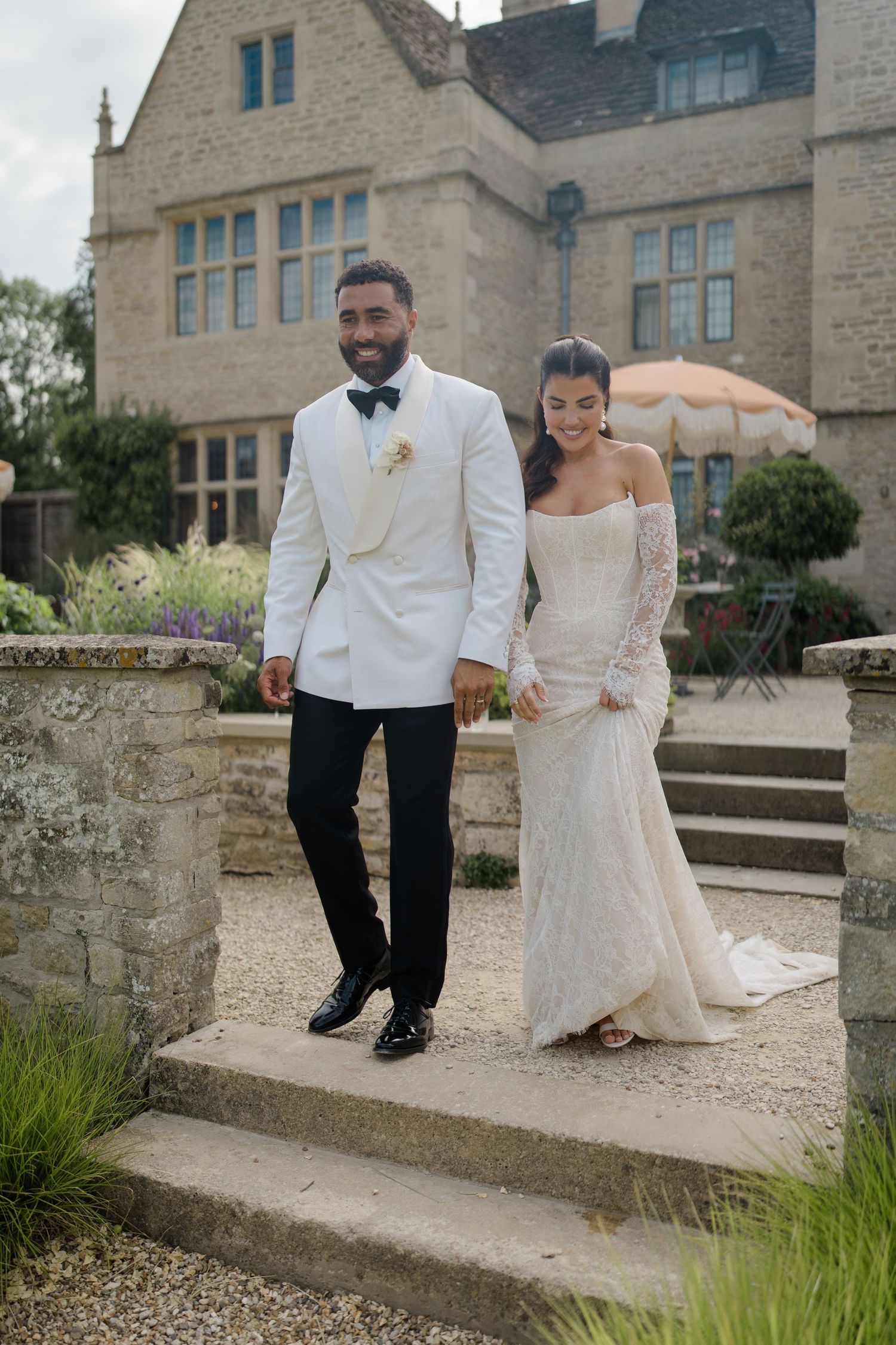 Newlywed couple posing on stone steps, groom in white tuxedo jacket and bride in elegant lace gown.