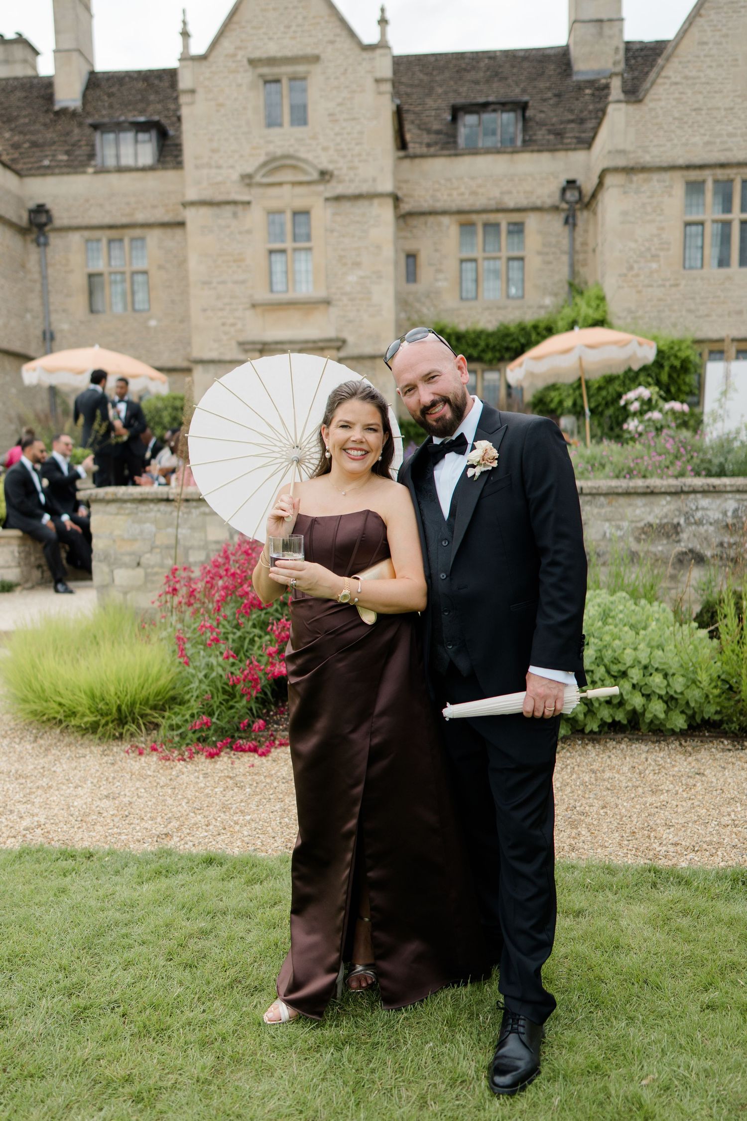 A couple in formal attire poses with a white parasol in front of a stone manor house.