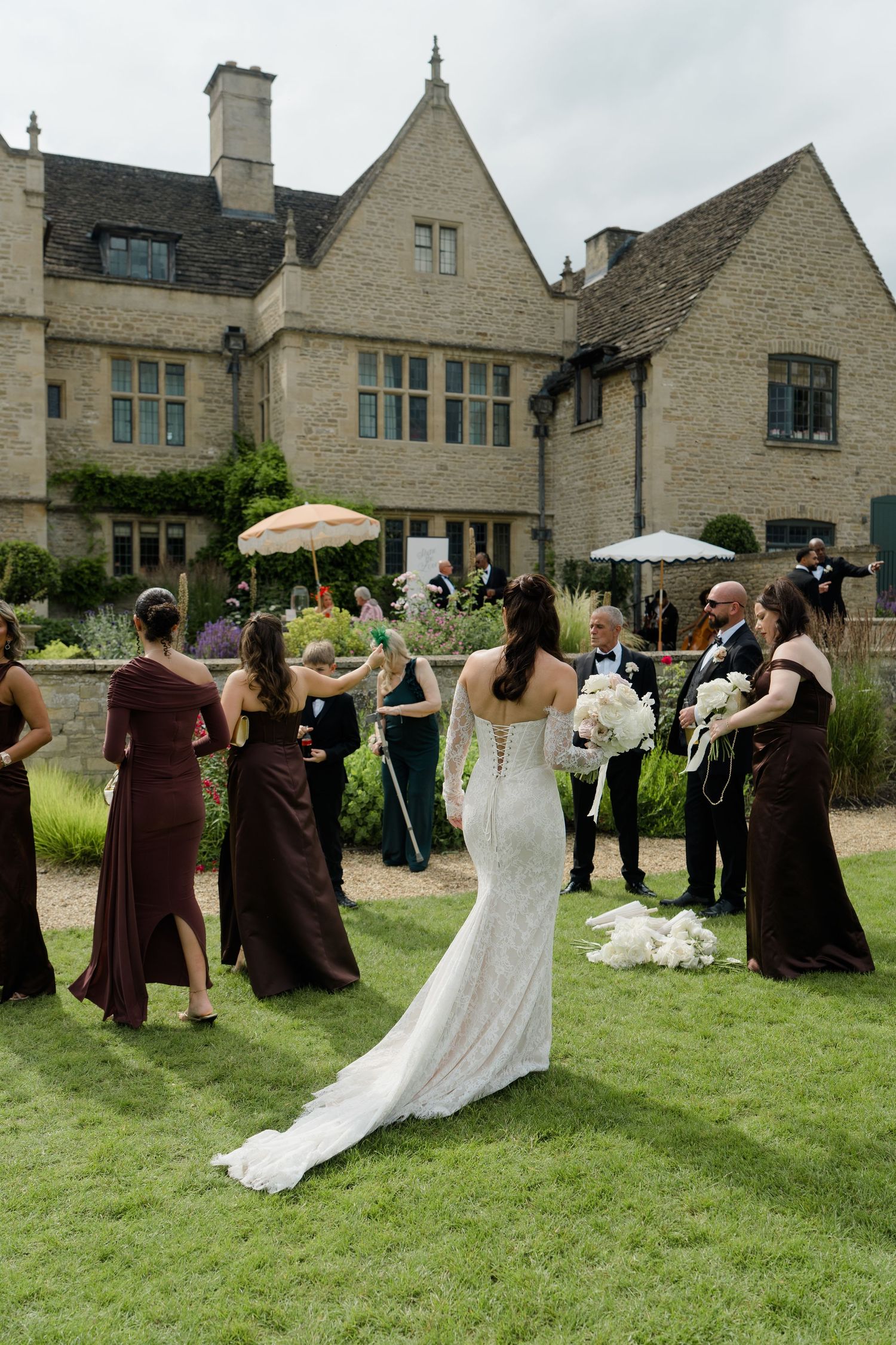 Bride in white fitted gown with train greeting bridesmaids in brown dresses outside manor.