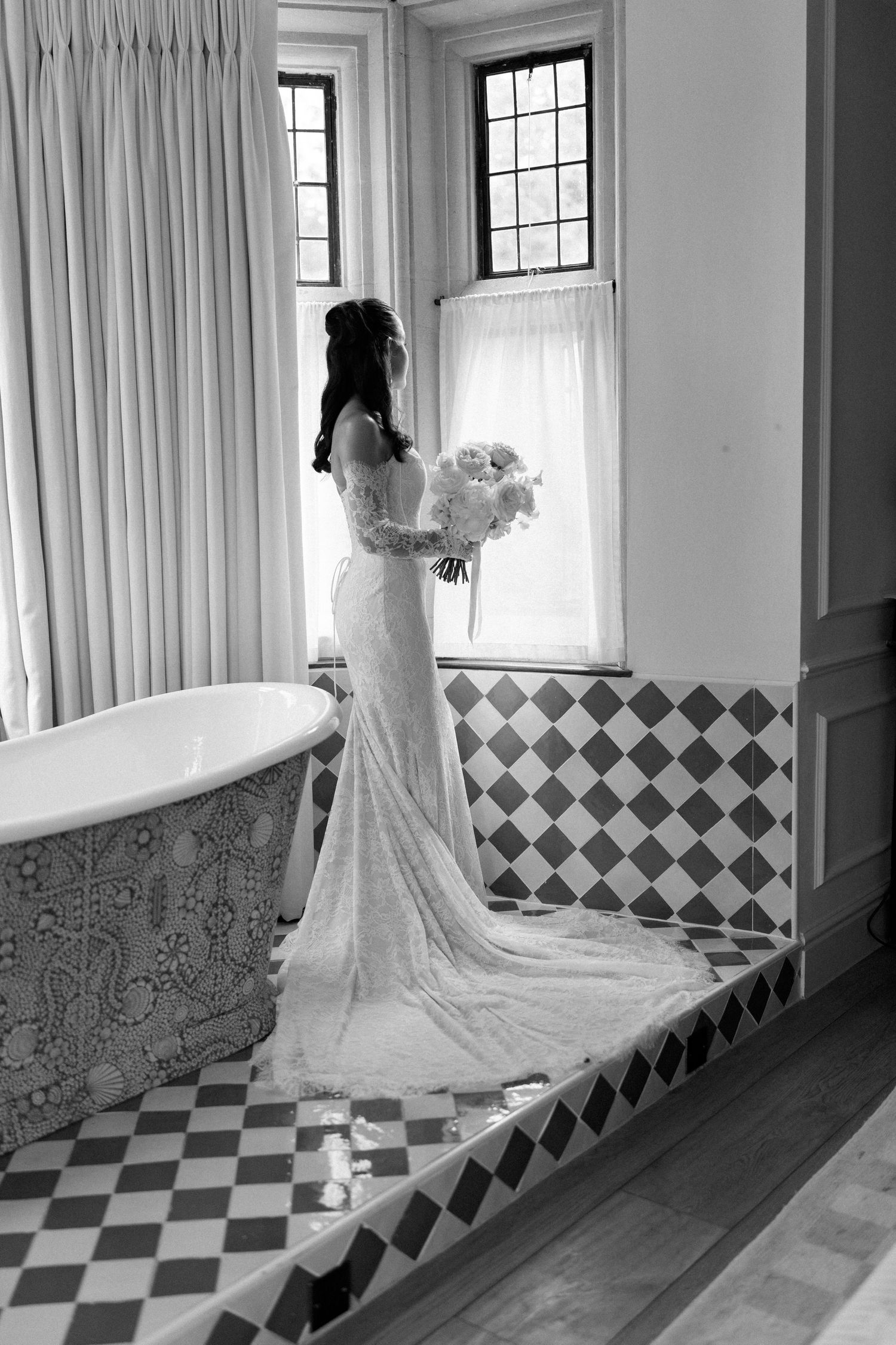 Bride in lace wedding gown holds white bouquet beside clawfoot tub in black and white photo.