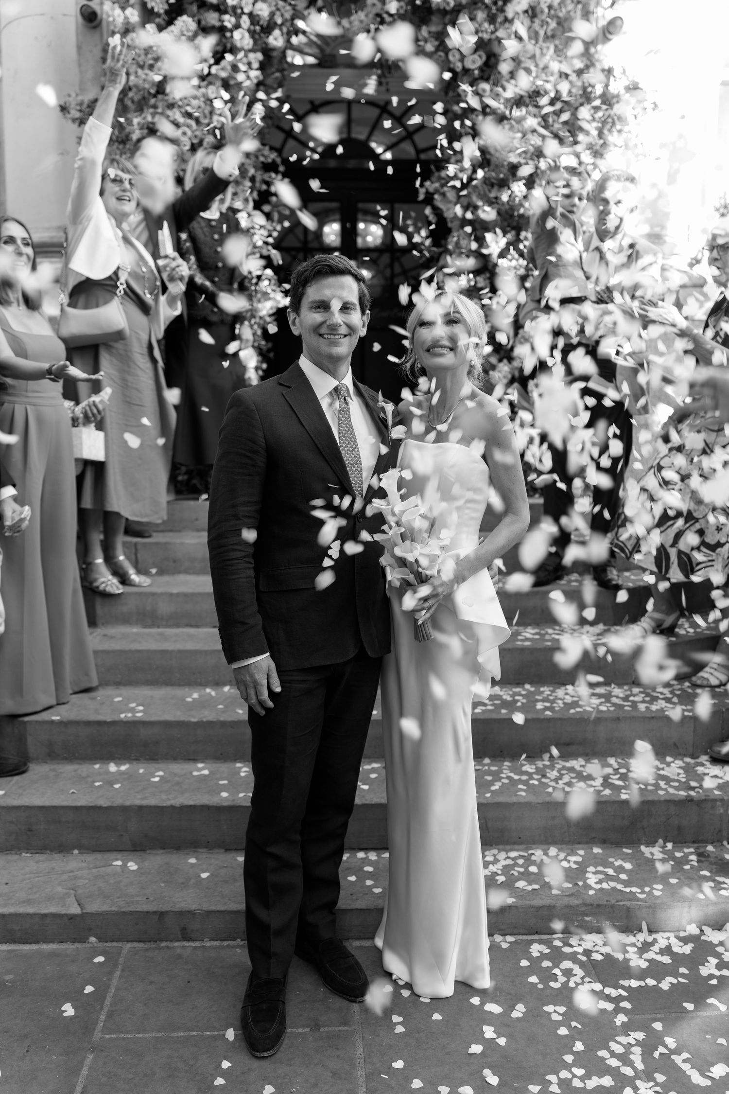 Black and white photo of a smiling bride and groom exiting church as guests toss flower petals in celebration.