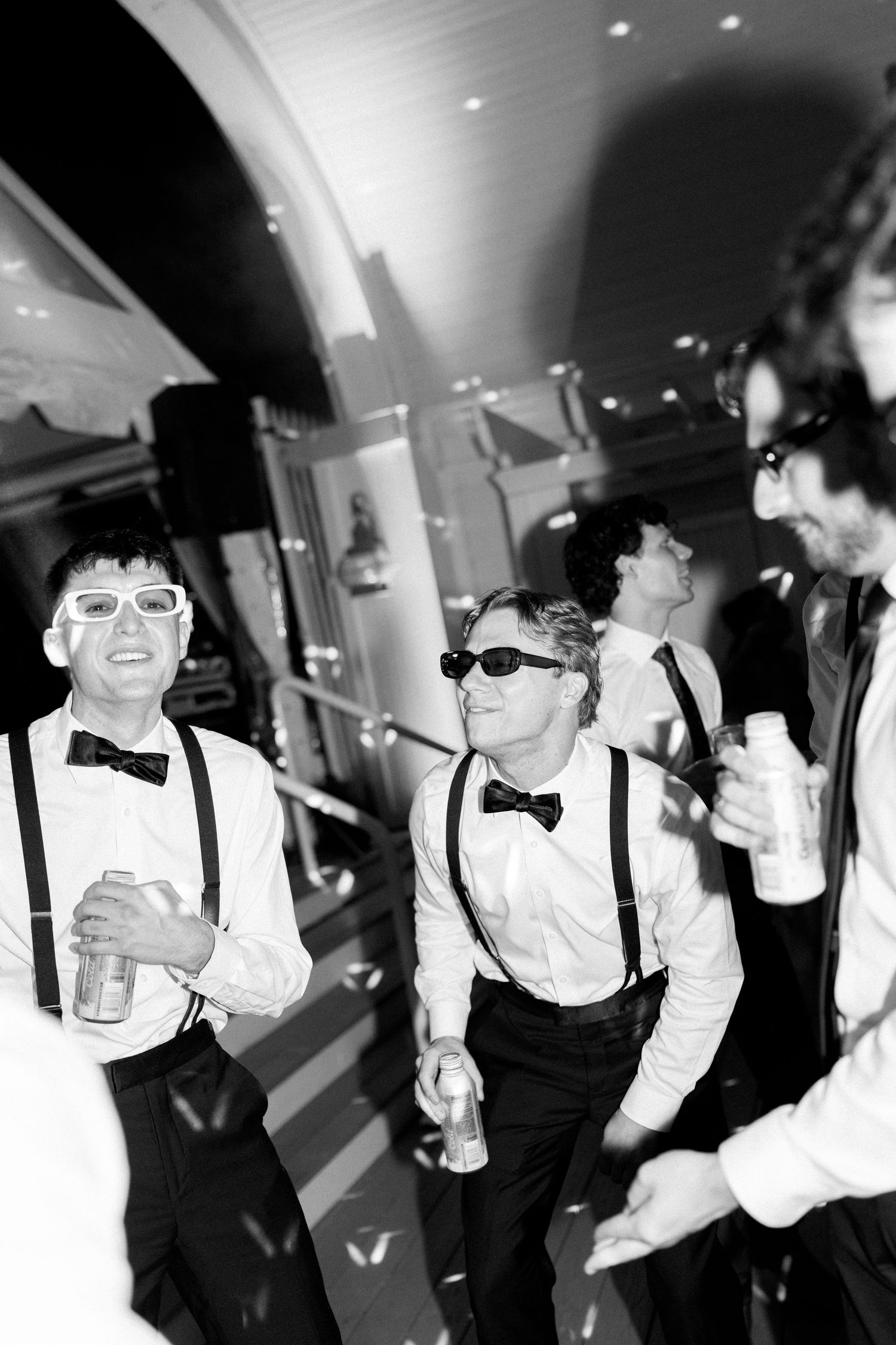 Black and white photo of groomsmen in suspenders and bow ties wearing sunglasses, holding drinks at a wedding reception.