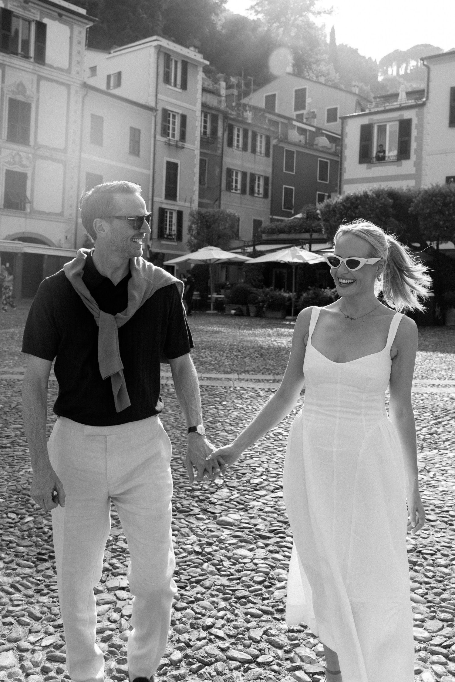 Black and white photo of a couple holding hands on cobblestone street in Italian village, woman in white dress and sunglasses.