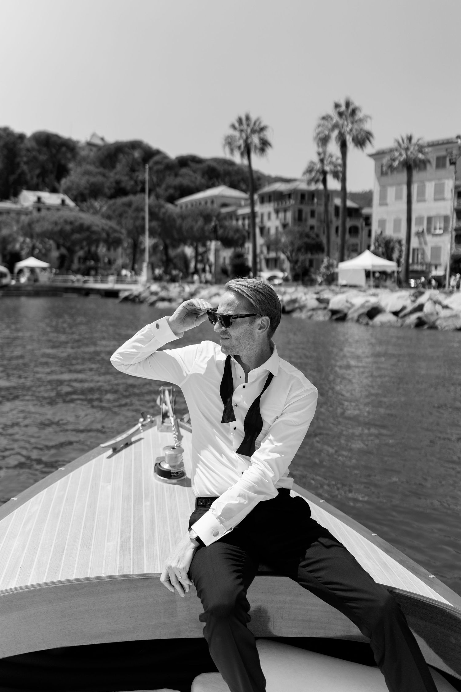 Stylish groom in white shirt and black tie sitting on bow of wooden boat in black and white photo.