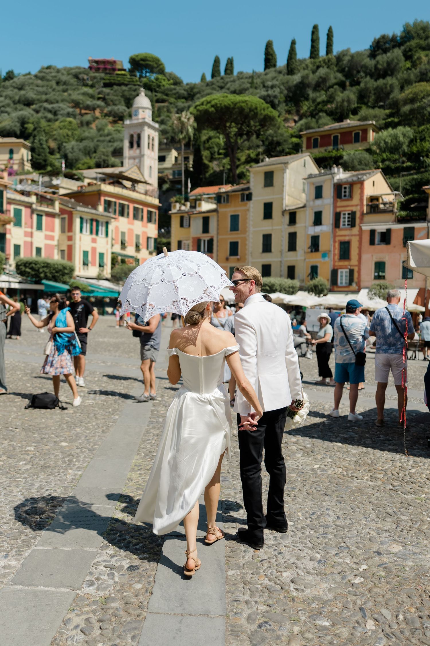 Newlyweds walk through colorful Portofino piazza, bride holding white parasol on sunny Italian wedding day.