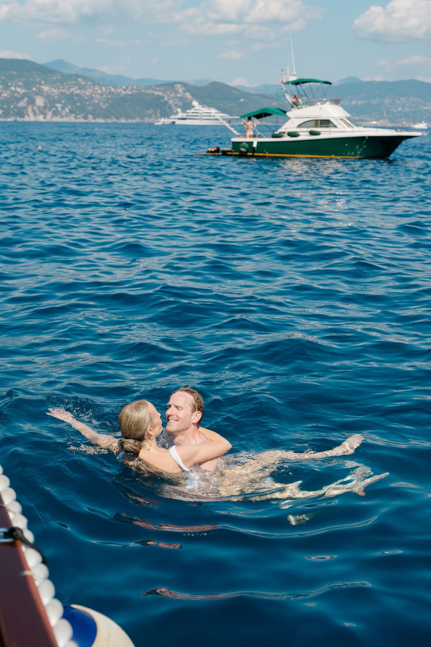 Happy couple swimming together in the blue Mediterranean Sea with yachts in the background.