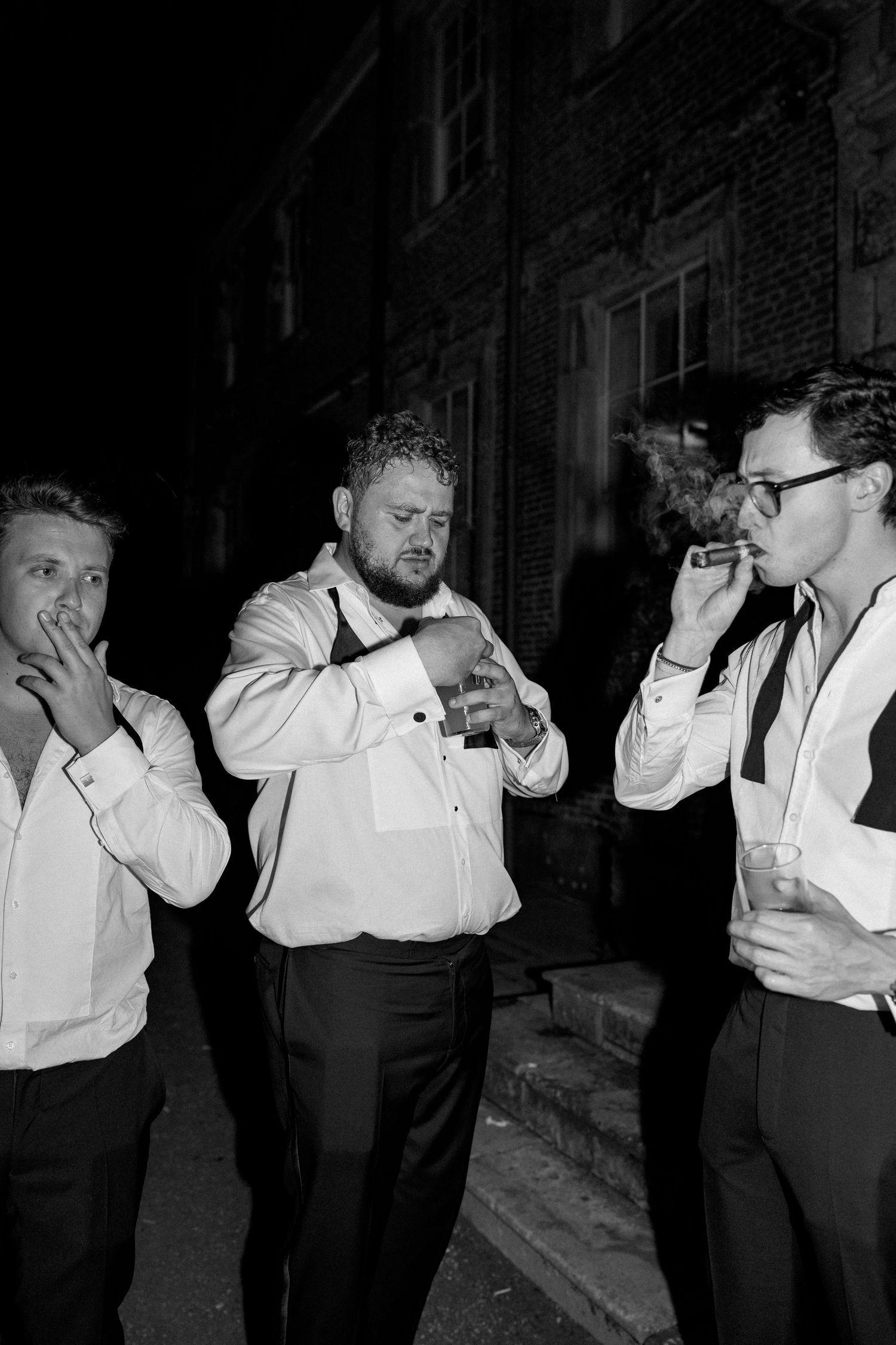 Black and white photo of three men smoking and drinking outside at a late-night wedding.